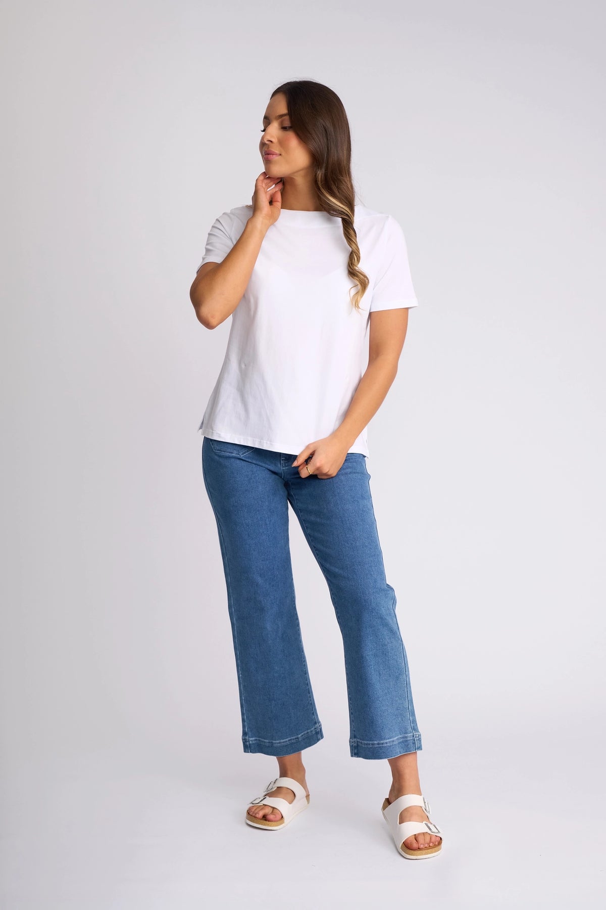 A person stands against a plain background wearing the Vassalli Raya Top in white, featuring a flattering boat neckline, paired with blue jeans and white sandals. The stretch cotton fabric of the Raya Top offers both comfort and style. They have long hair, with one hand touching their chin and the other resting by their side.