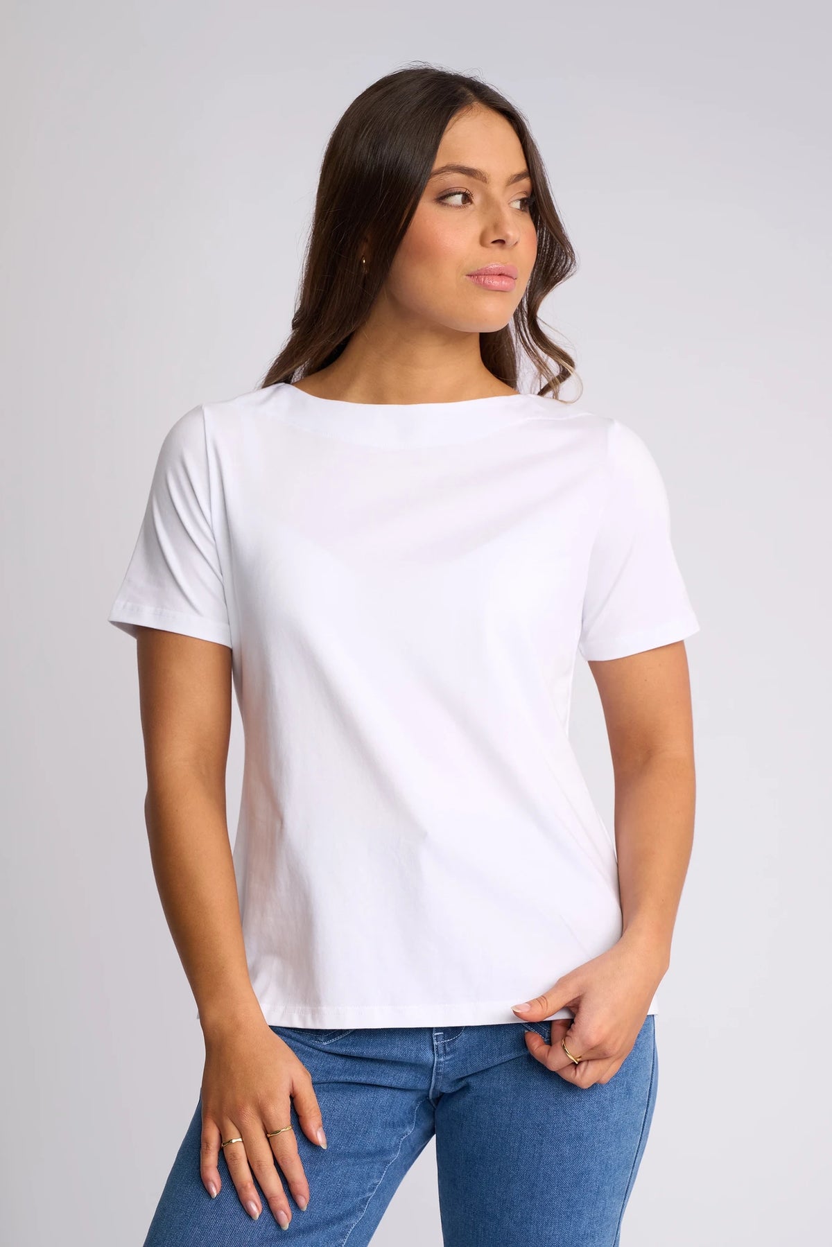 A woman with long brown hair is wearing the Vassalli Raya Top in white, featuring a boat neckline and short sleeves in soft stretch cotton. She pairs it with blue jeans and stands against a light gray background, looking slightly to the side.