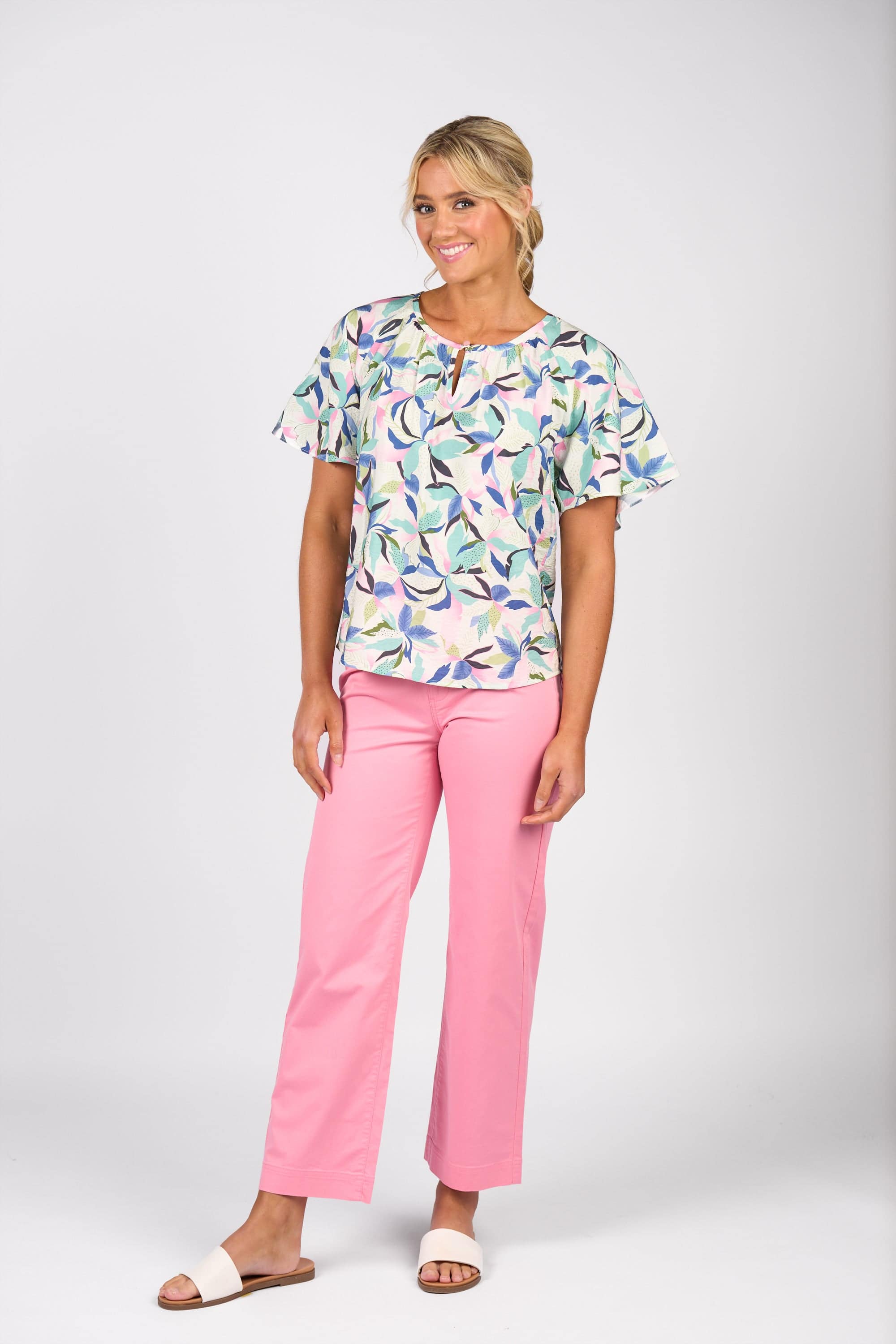 A woman stands against a white background, smiling with her blonde hair tied back. She wears the Vassalli Floaty Sleeve Top - Soleil, pink pants, and white sandals.