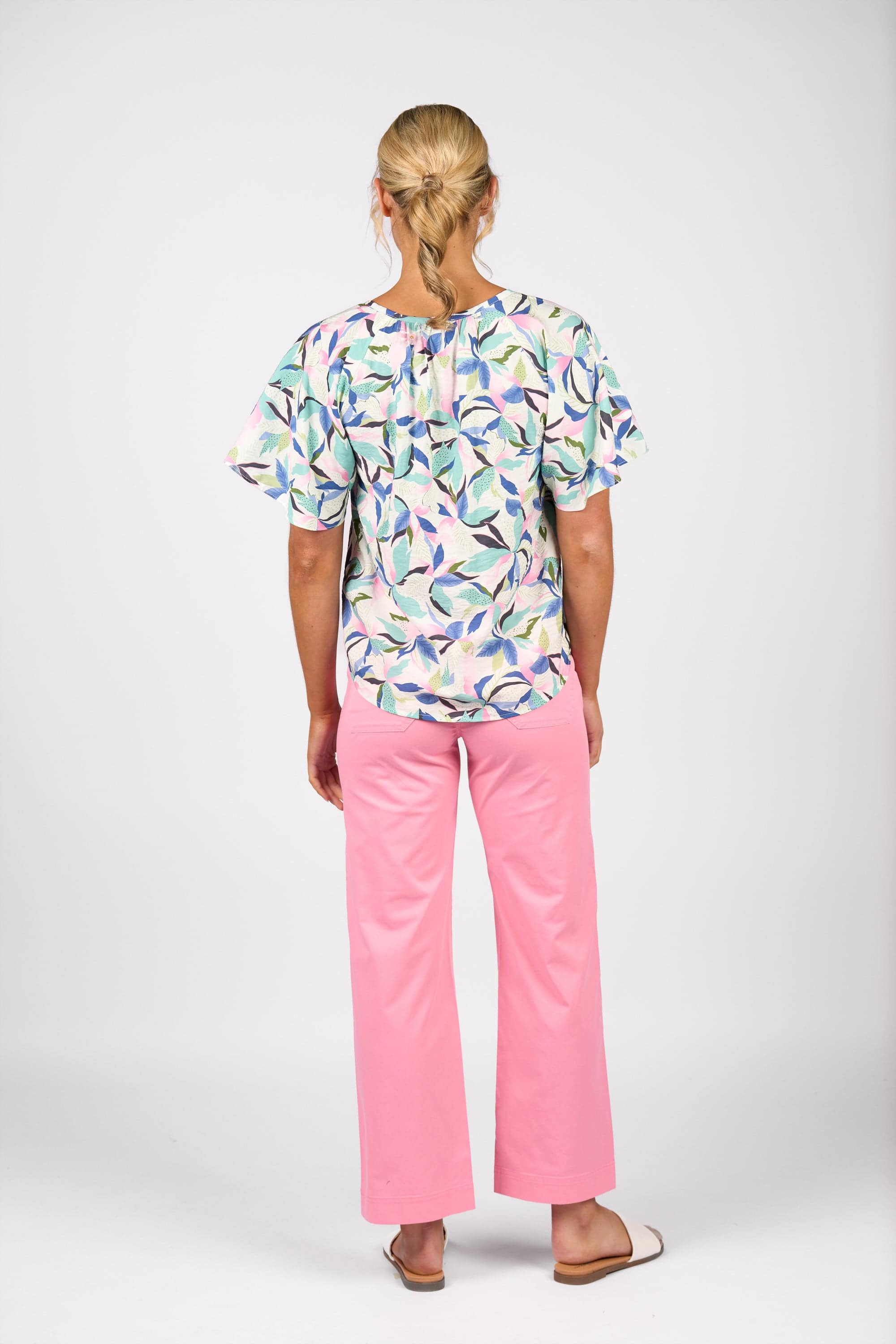 A person with blonde hair in a low bun, seen from behind, wears the Vassalli Floaty Sleeve Top - Soleil with pink pants and white sandals, standing against a plain white background.