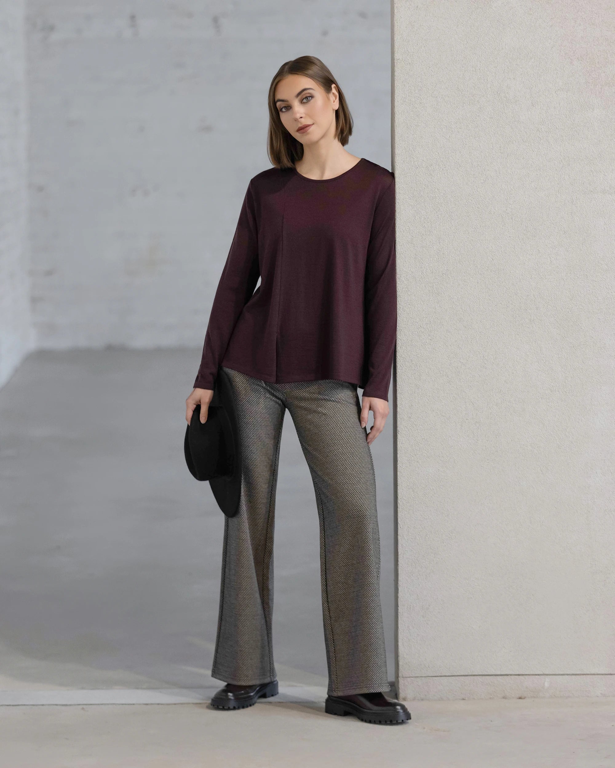 A woman stands indoors against a light wall, wearing the Vassalli Pave Merino Top in Berry with gray wide-leg pants and black shoes. She holds a black hat in her left hand and looks at the camera.