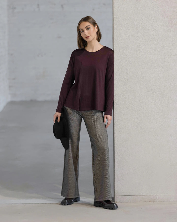 A woman stands indoors against a light wall, wearing the Vassalli Pave Merino Top in Berry with gray wide-leg pants and black shoes. She holds a black hat in her left hand and looks at the camera.