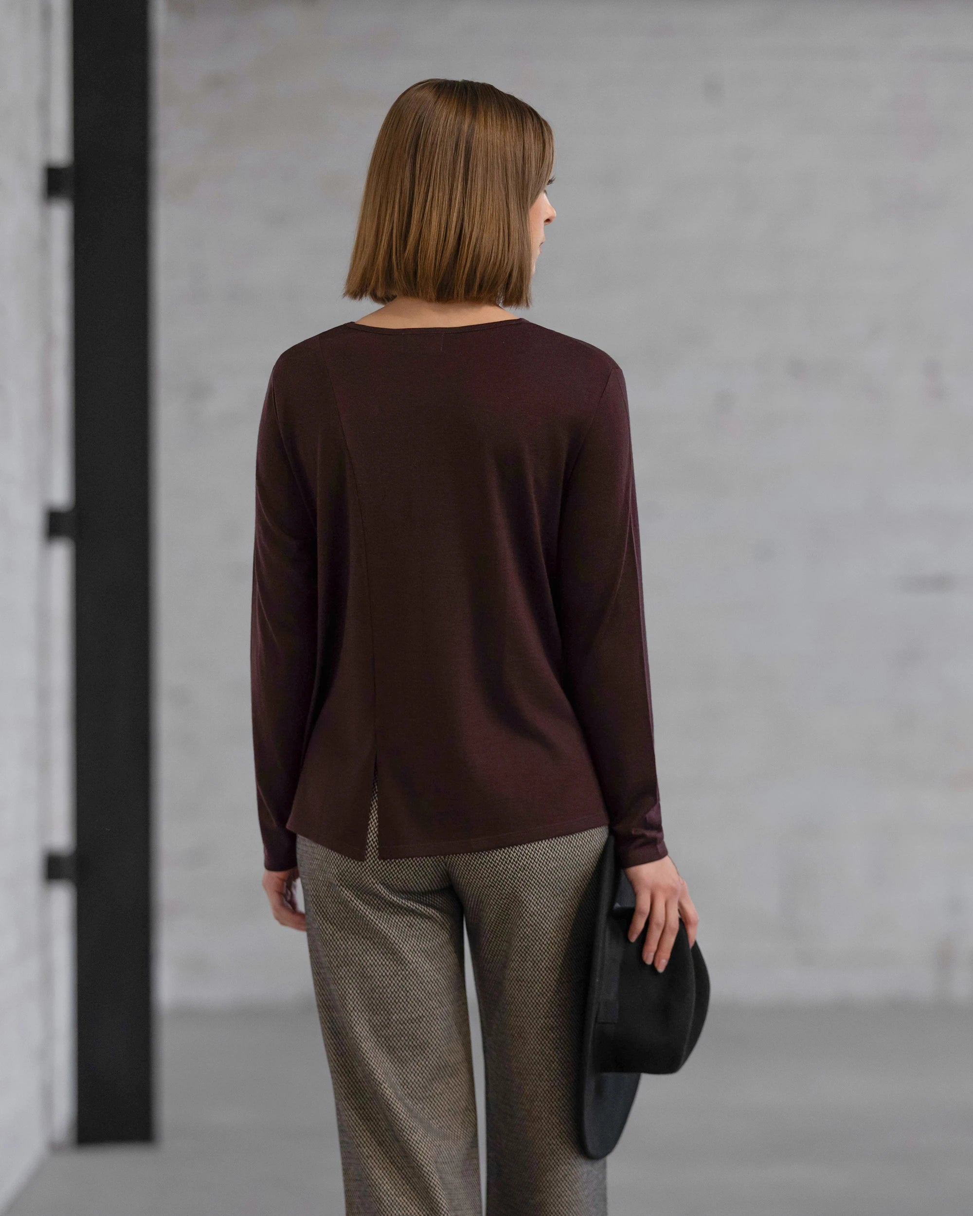 A woman with straight, shoulder-length hair faces away indoors, wearing the Vassalli Pave Merino Top in Berry with gray pants and holding a black hat by her side against a plain, light brick wall.
