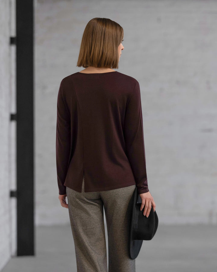 A woman with straight, shoulder-length hair faces away indoors, wearing the Vassalli Pave Merino Top in Berry with gray pants and holding a black hat by her side against a plain, light brick wall.