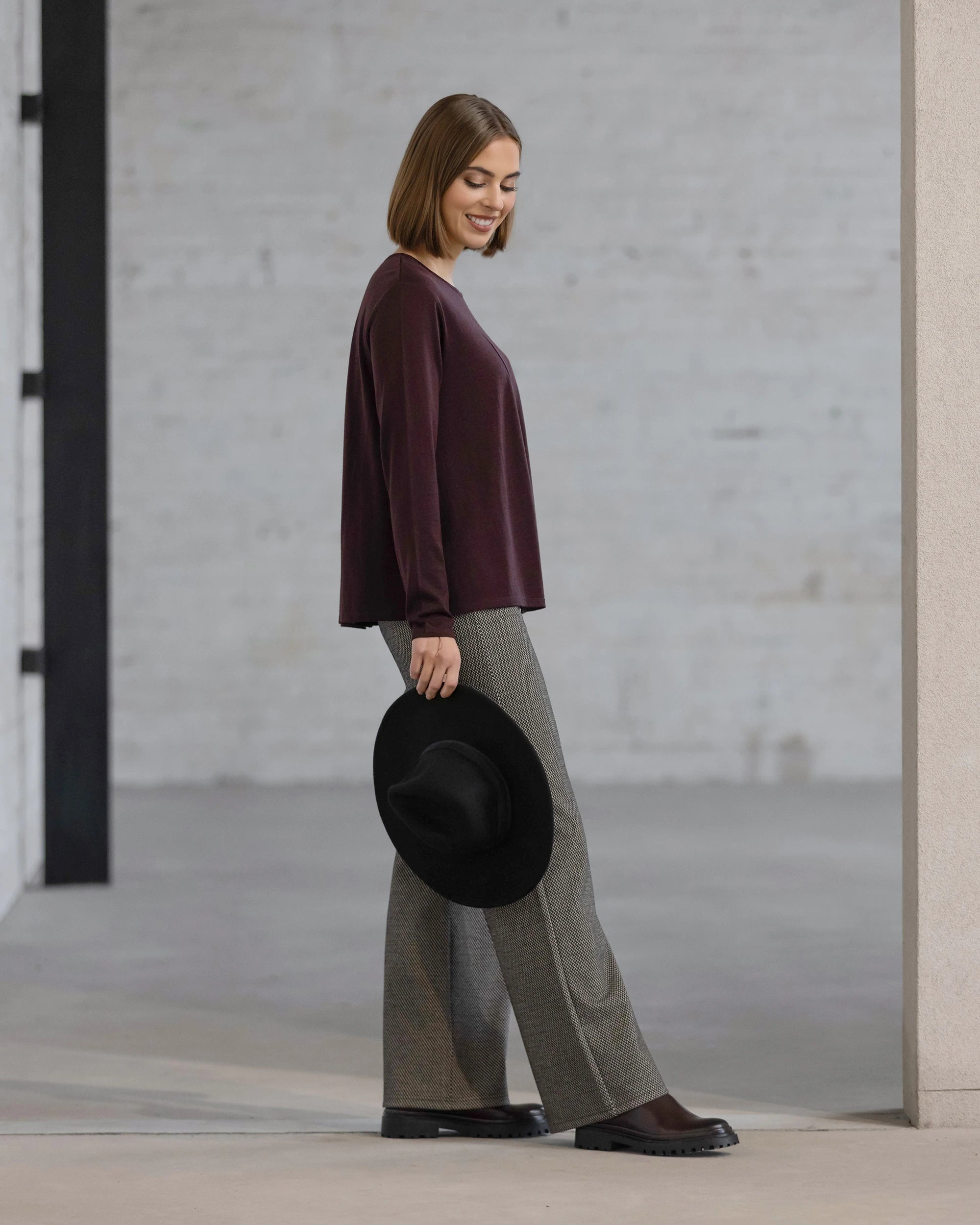 A woman with straight brown hair wears the Vassalli Pave Merino Top in Berry and gray wide-leg pants, holding a black hat and smiling against a minimalist background with light gray brick walls.