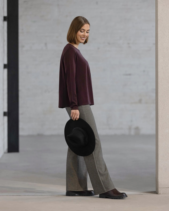 A woman with straight brown hair wears the Vassalli Pave Merino Top in Berry and gray wide-leg pants, holding a black hat and smiling against a minimalist background with light gray brick walls.