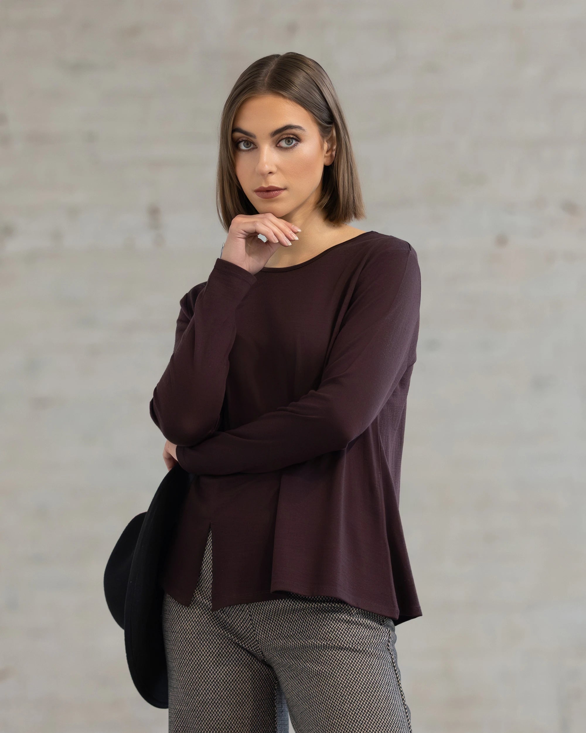 A woman with straight brown hair and light makeup poses indoors against a light brick wall, wearing the Vassalli Pave Merino Top in Berry with patterned pants, holding a black hat at her side and resting her chin on her hand.