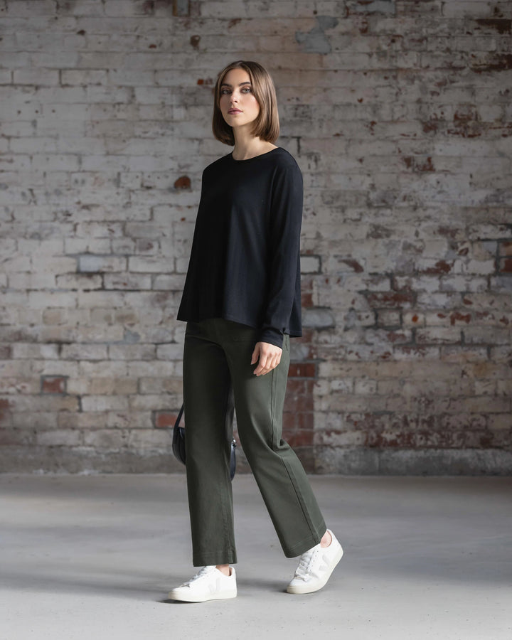 A woman wearing the Vassalli Pave Merino Top in black stands before a weathered brick wall, paired with green pants and white sneakers, gazing neutrally at the camera.