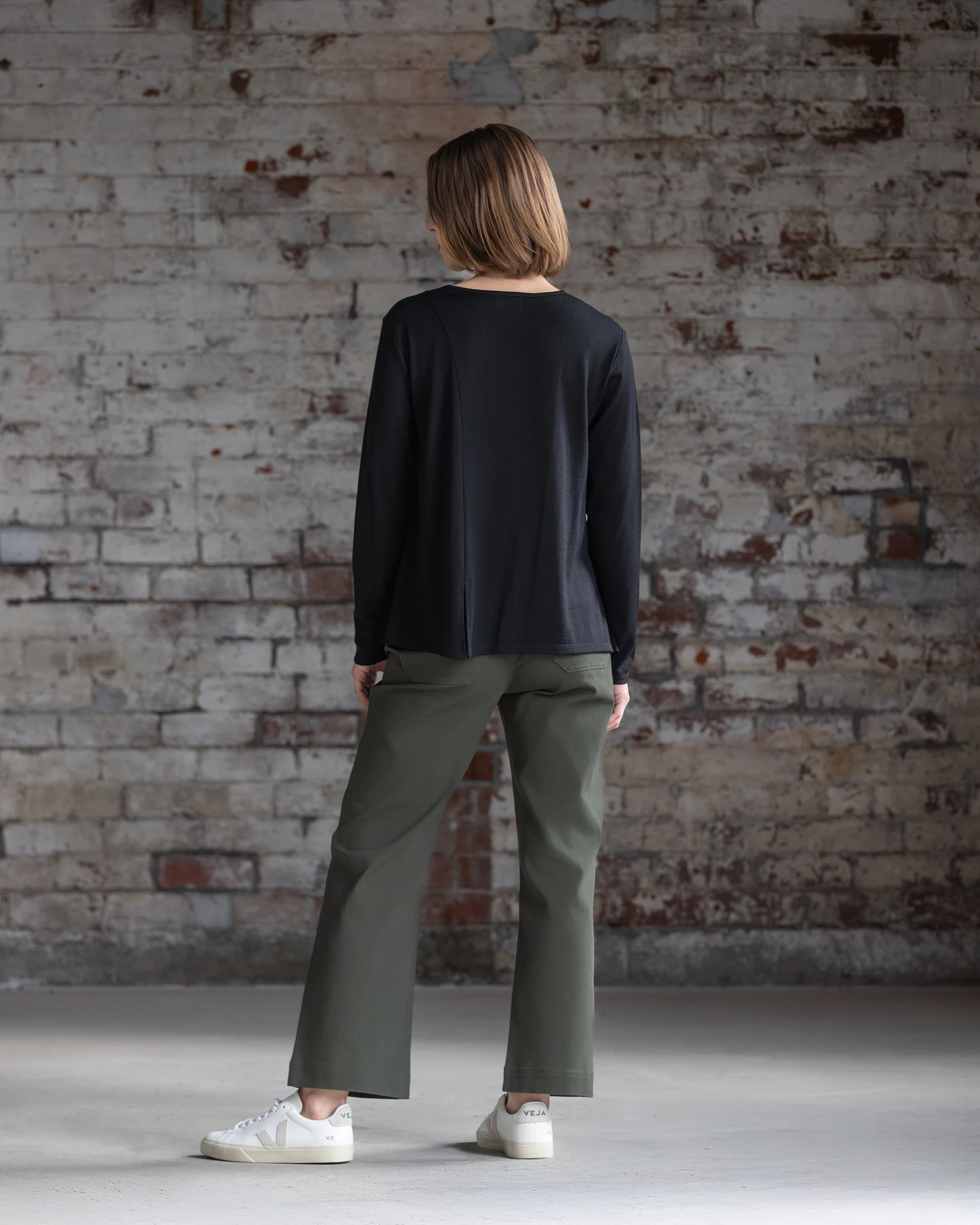 A person with short light brown hair, dressed in the Vassalli Pave Merino Top in black, olive green pants, and white sneakers, stands indoors facing a weathered brick wall.