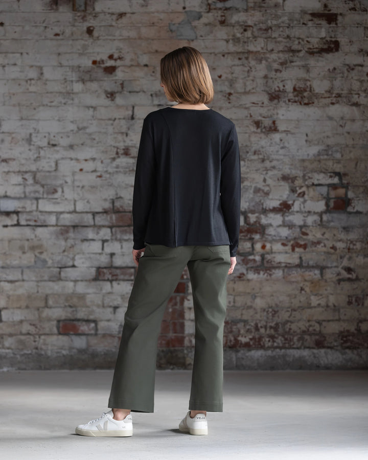 A person with short light brown hair, dressed in the Vassalli Pave Merino Top in black, olive green pants, and white sneakers, stands indoors facing a weathered brick wall.