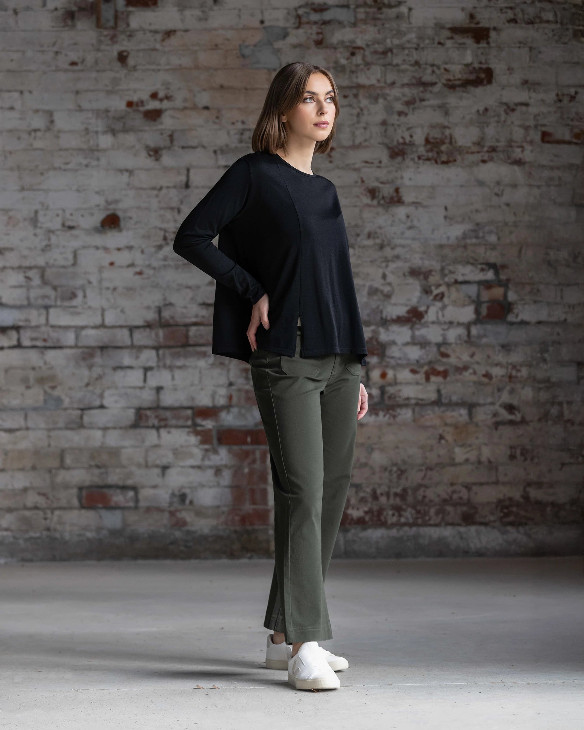 A woman stands indoors before a textured brick wall, wearing the Vassalli Pave Merino Top in Black, olive green pants, and white shoes. She looks to the side with one hand on her hip.