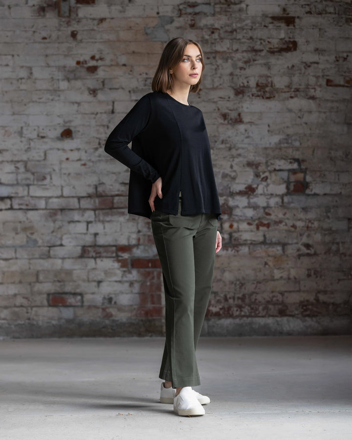 A woman stands indoors before a textured brick wall, wearing the Vassalli Pave Merino Top in Black, olive green pants, and white shoes. She looks to the side with one hand on her hip.