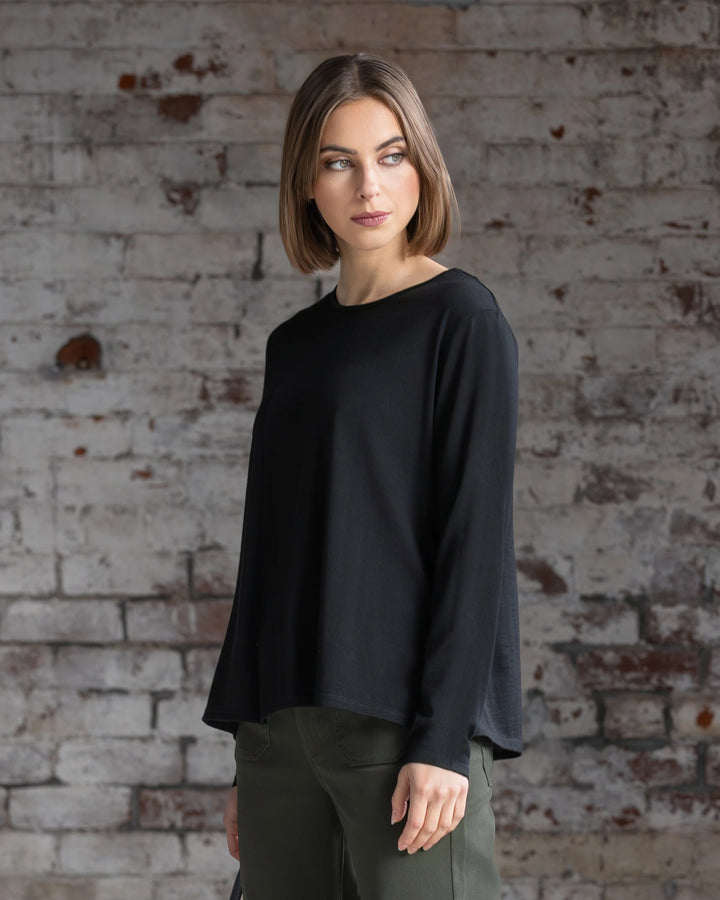 A woman with a bob haircut, wearing the Vassalli Pave Merino Top in black and green pants, stands before a weathered brick wall, gazing to the side.