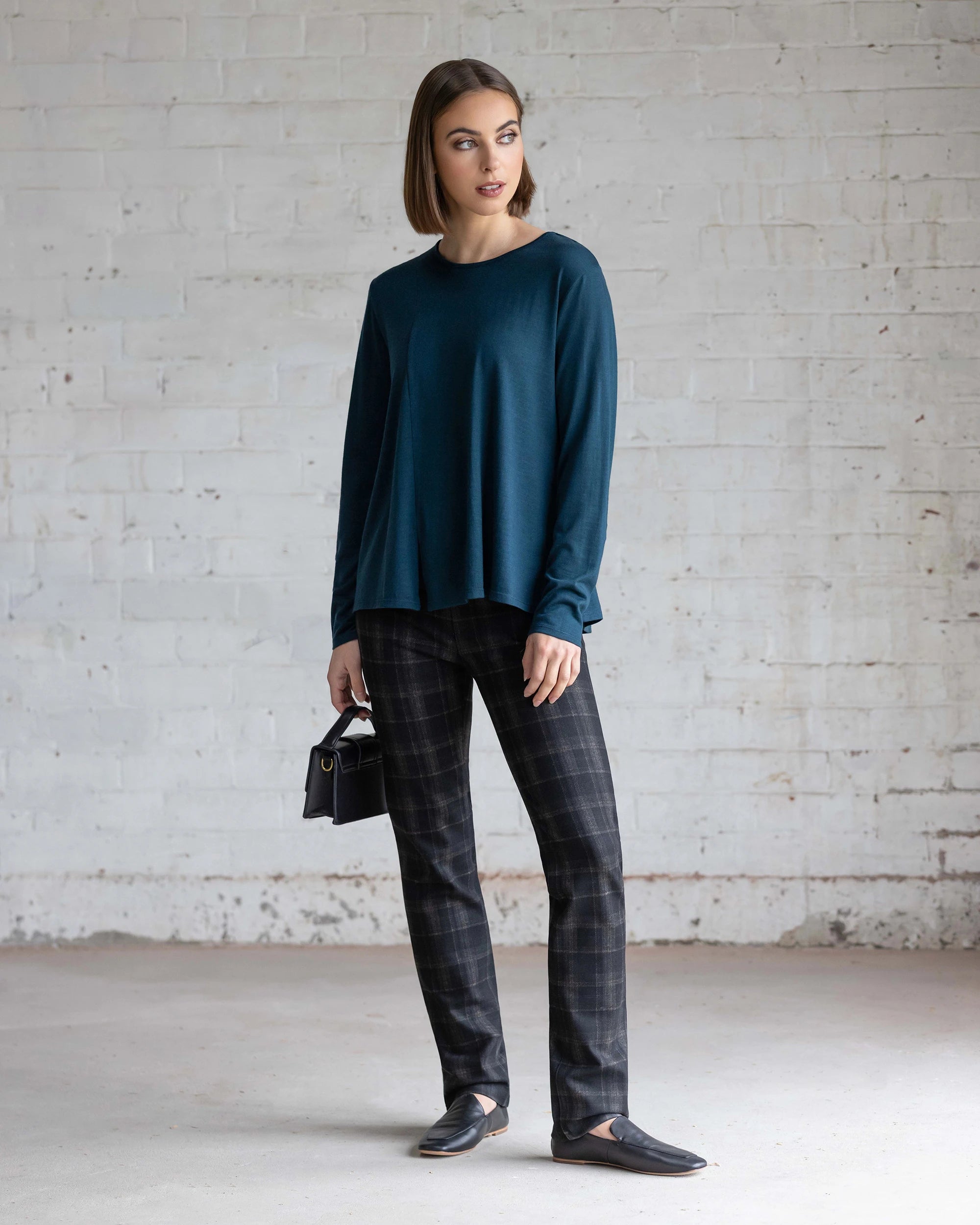 A woman stands indoors against a white brick wall, wearing the Vassalli Pave Merino Top in Dragonfly with black plaid pants, black loafers, and a small black handbag. She has straight, shoulder-length hair and a neutral expression.