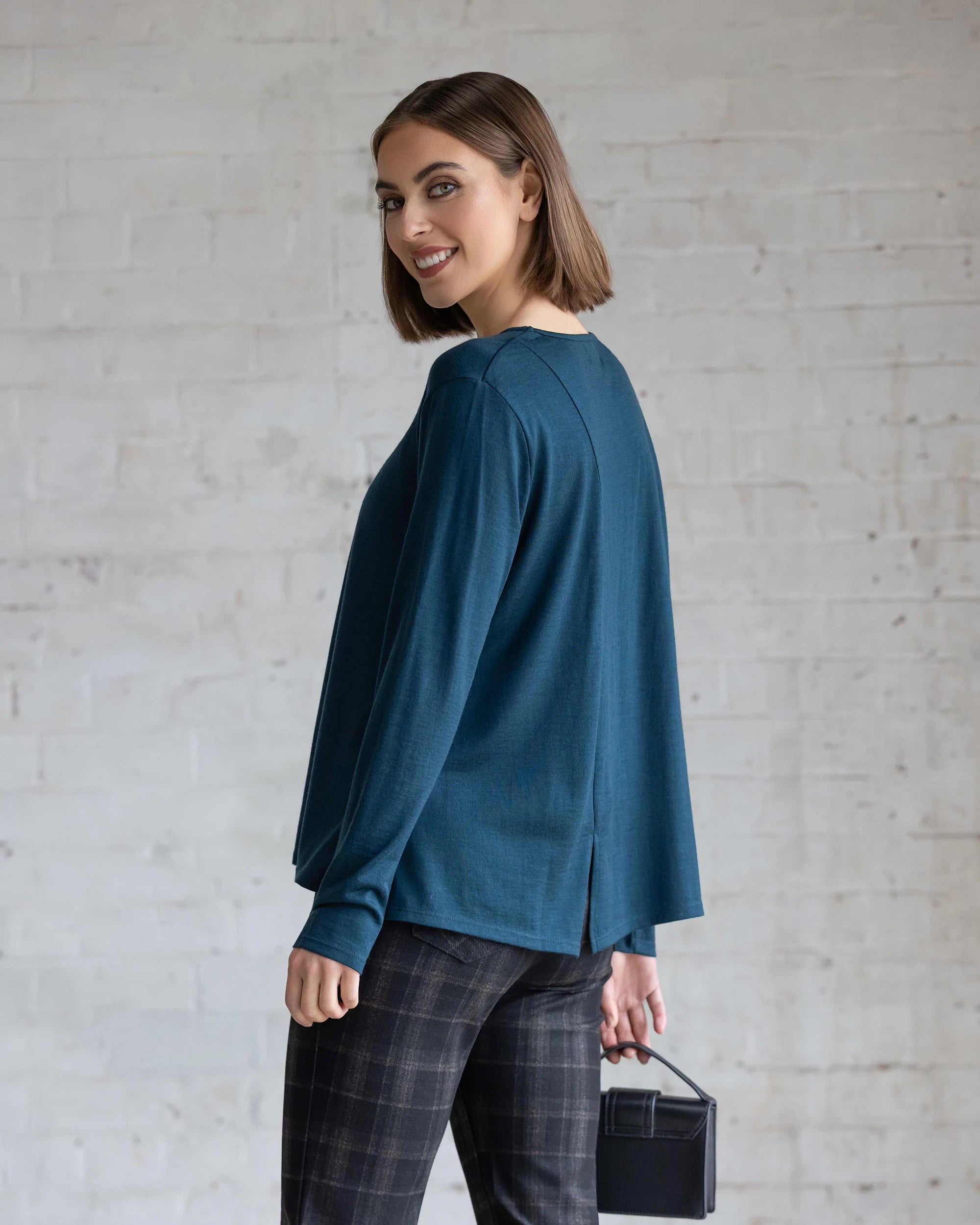 A woman with a chin-length bob smiles over her shoulder, wearing the Vassalli Pave Merino Top in Dragonfly, plaid pants, and holding a small black box bag against a light brick wall.