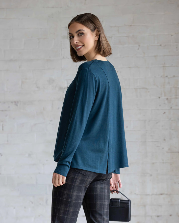 A woman with a chin-length bob smiles over her shoulder, wearing the Vassalli Pave Merino Top in Dragonfly, plaid pants, and holding a small black box bag against a light brick wall.