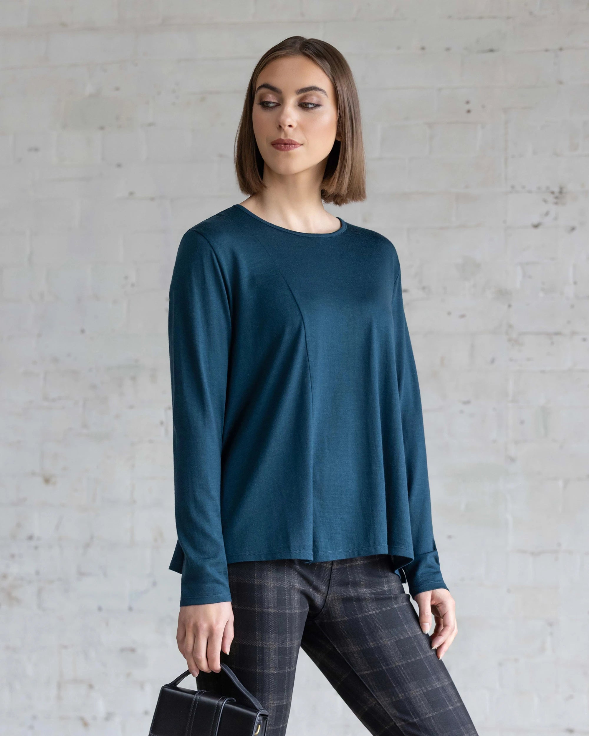 A woman with straight brown hair wears the Vassalli Pave Merino Top in Dragonfly and plaid pants, standing before a light-colored brick wall, holding a small black handbag and gazing slightly to the side.
