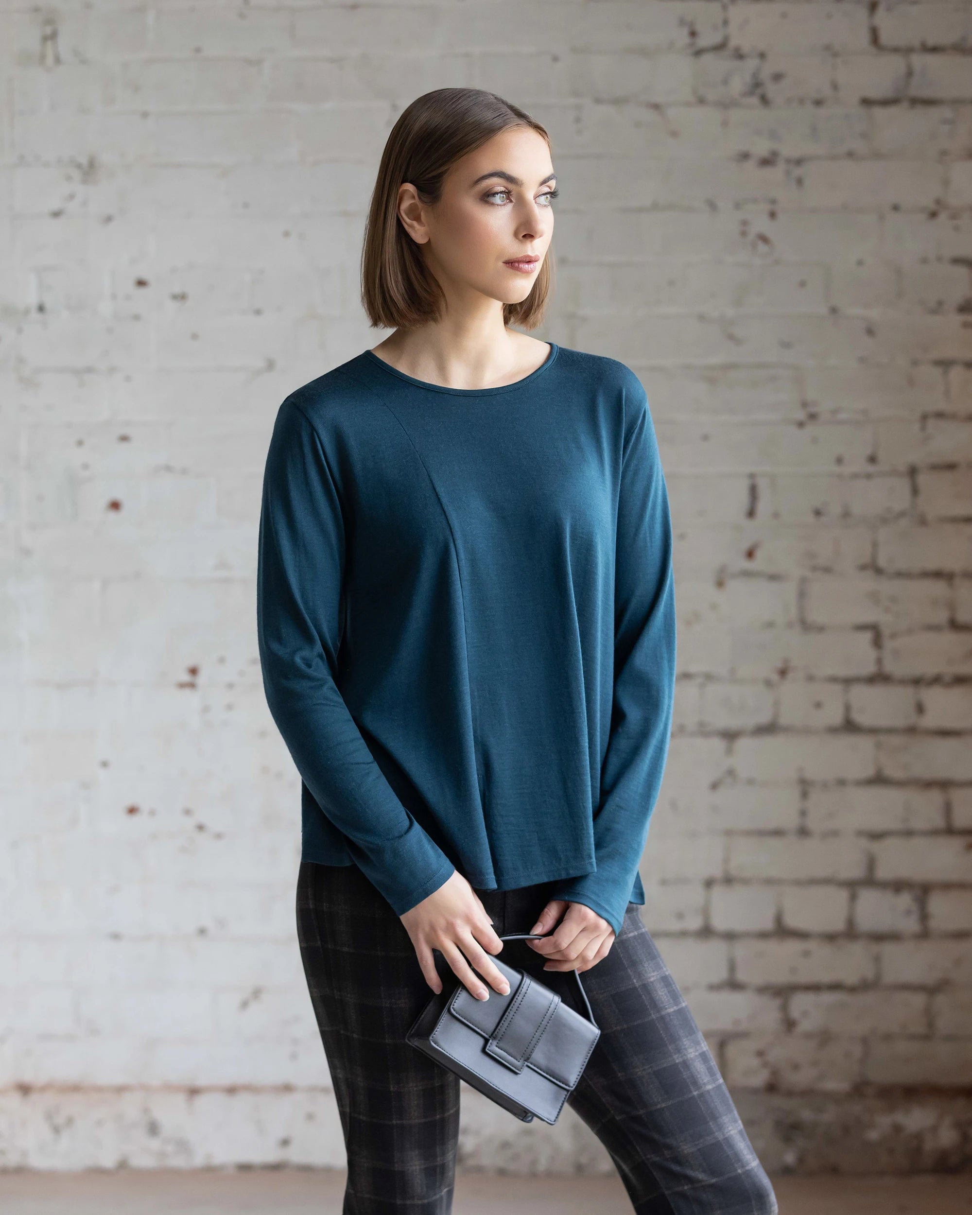A woman wears the Vassalli Pave Merino Top in Dragonfly, styled with dark grey plaid trousers and a small grey structured bag, standing against a white brick wall.