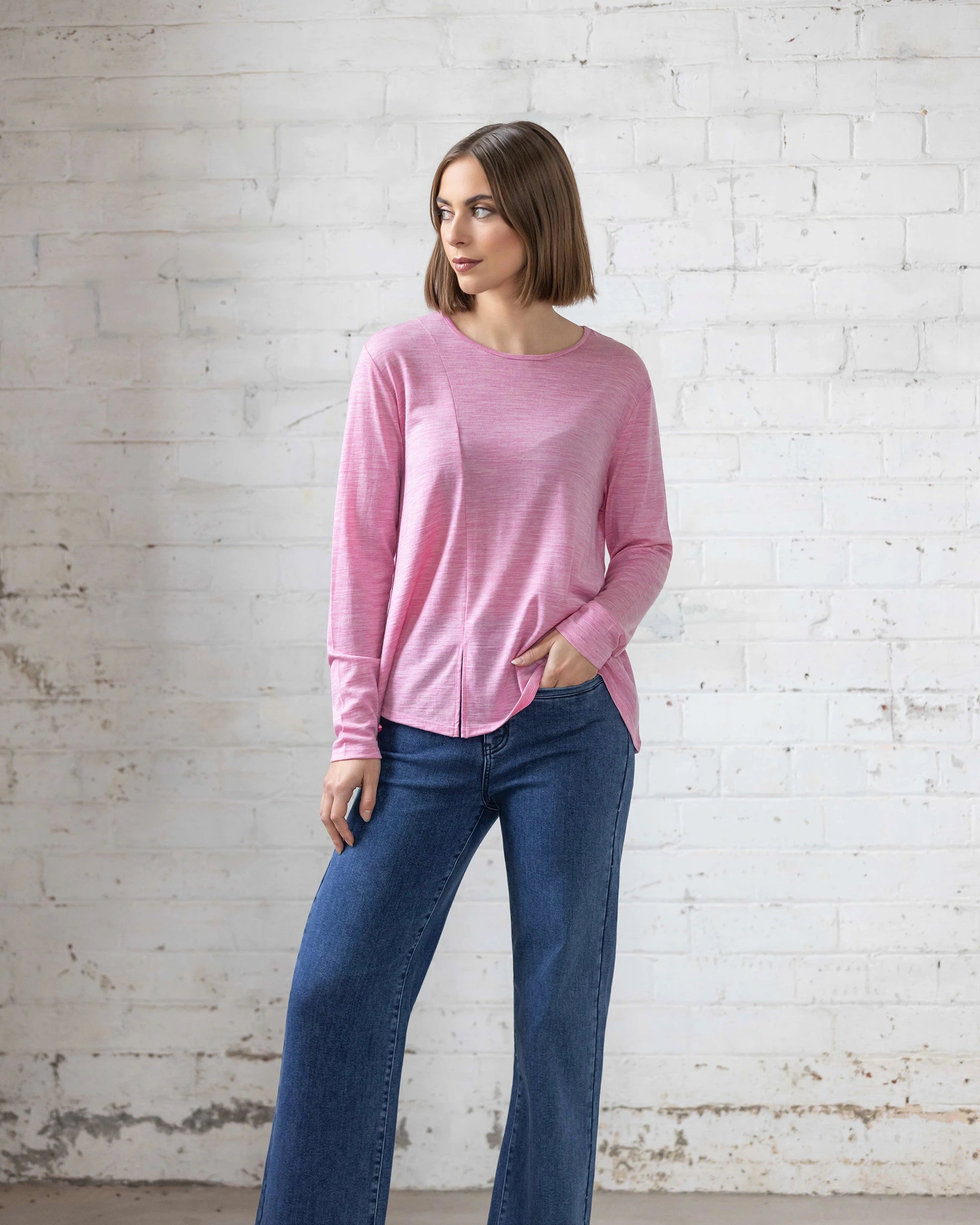 A woman with a bob haircut models the Vassalli Pave Merino Top in Pink Marle and wide-leg blue jeans, standing against a white brick wall with one hand in her pocket—ideal for winter layering.