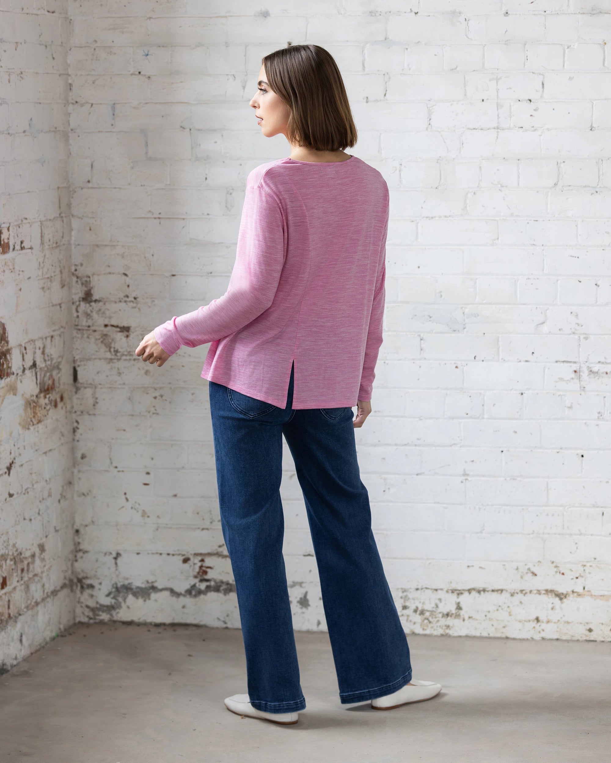 Indoors against a white brick wall, someone wears the Vassalli Pave Merino Top in Pink Marle, styled with blue wide-leg jeans and white shoes. The minimalist setting highlights this cozy, versatile piece—ideal for winter layering.