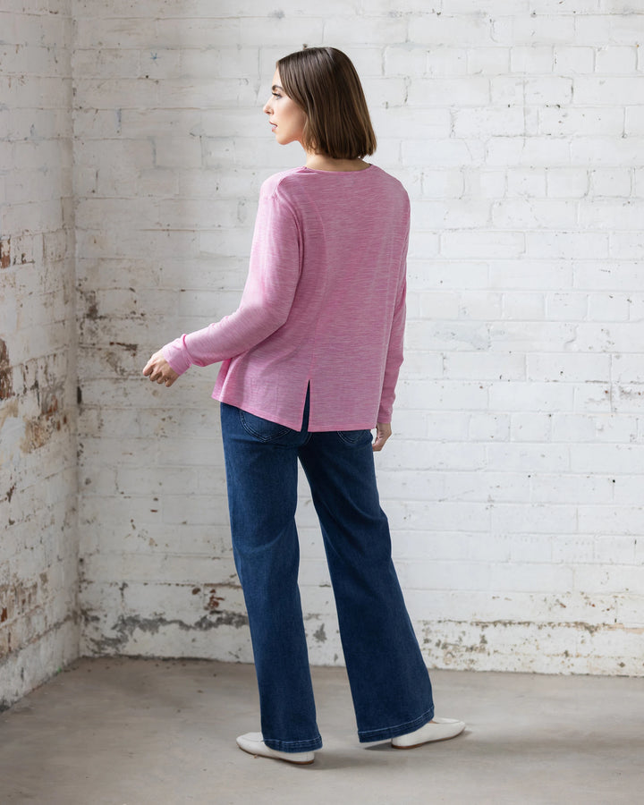 Indoors against a white brick wall, someone wears the Vassalli Pave Merino Top in Pink Marle, styled with blue wide-leg jeans and white shoes. The minimalist setting highlights this cozy, versatile piece—ideal for winter layering.