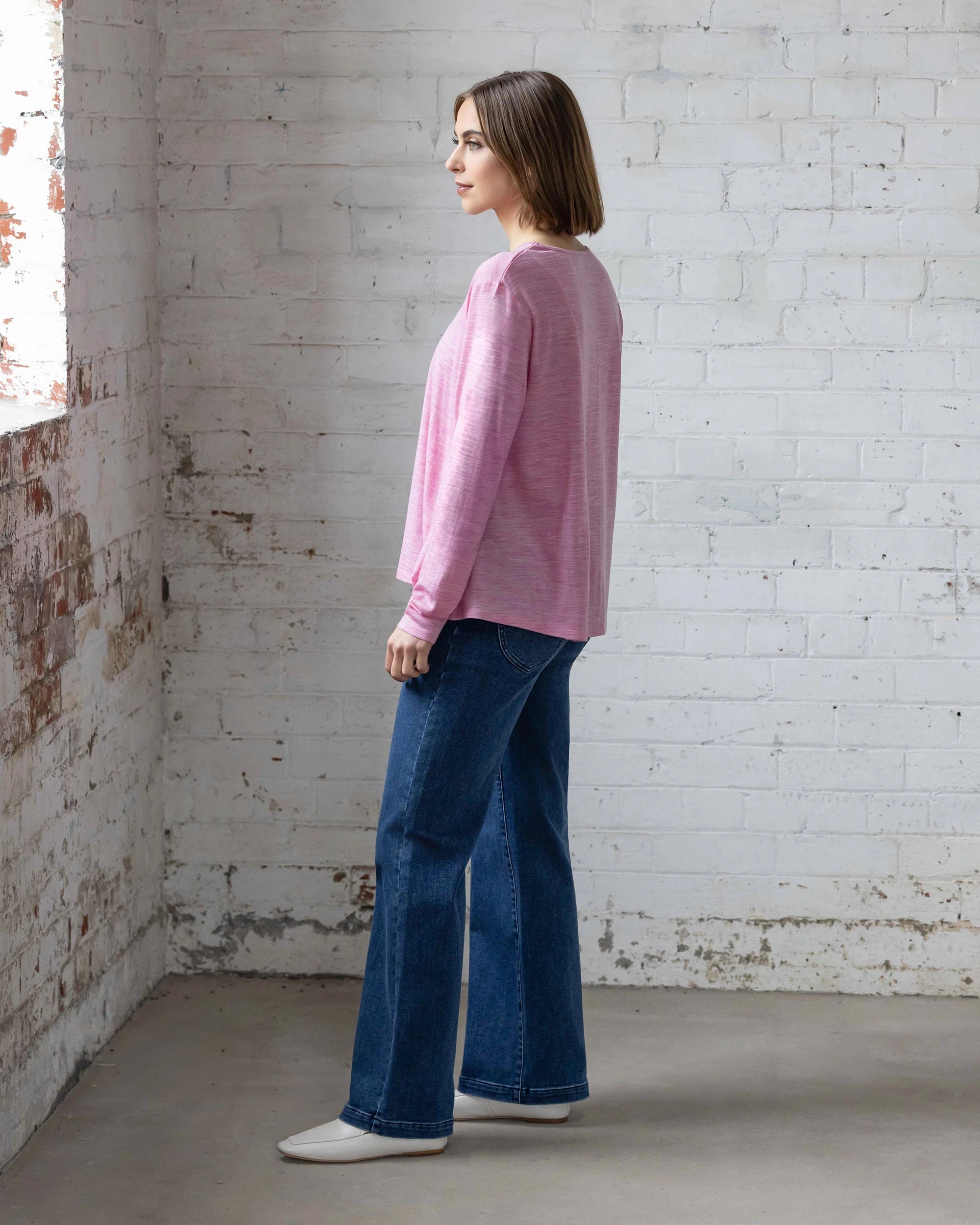 A woman with straight brown hair stands sideways in a sunlit room, wearing the Vassalli Pave Merino Top in Pink Marle, blue jeans, and white shoes. She faces a window set in a white brick wall—perfect for winter layering.