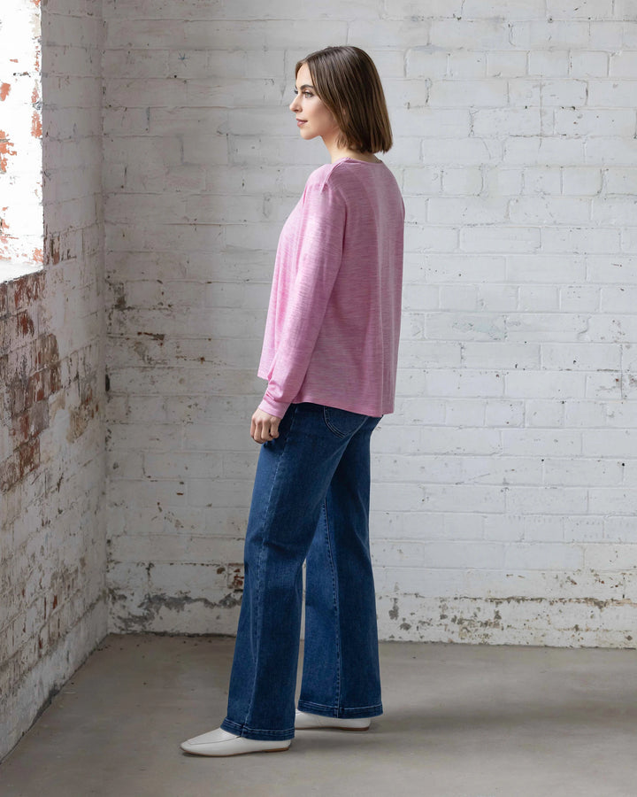 A woman with straight brown hair stands sideways in a sunlit room, wearing the Vassalli Pave Merino Top in Pink Marle, blue jeans, and white shoes. She faces a window set in a white brick wall—perfect for winter layering.