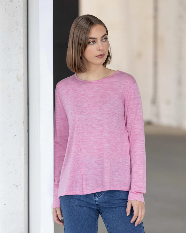 A woman with straight, shoulder-length brown hair wears the Vassalli Pave Merino Top in Pink Marle and blue jeans. She stands indoors near a white wall, looking to her left—ideal for winter layering.