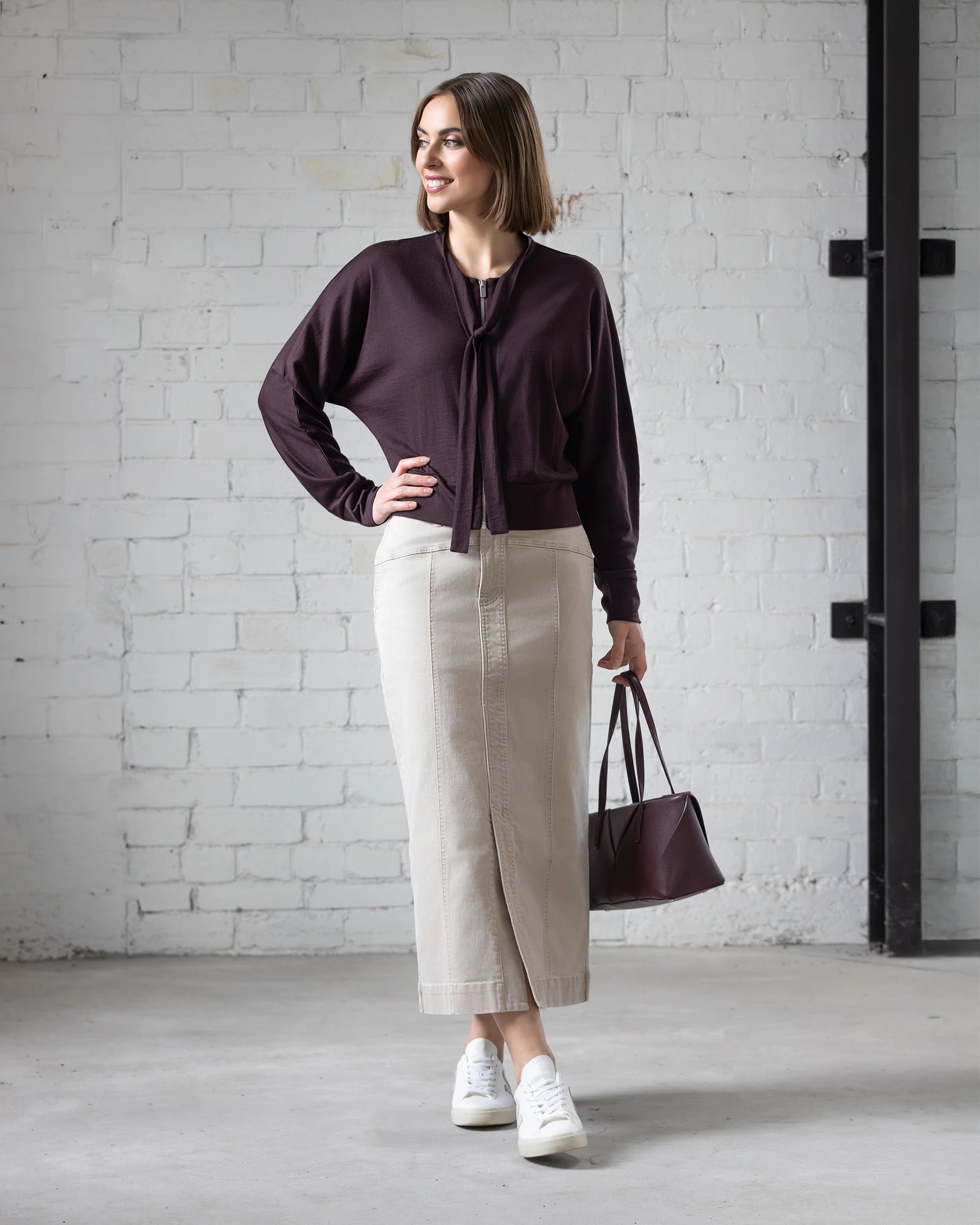 A woman stands indoors against a white brick wall, wearing the Vassalli Nina Merino Cardi in Berry over a beige ankle-length skirt with white sneakers. She carries a dark handbag and smiles while looking to the side.