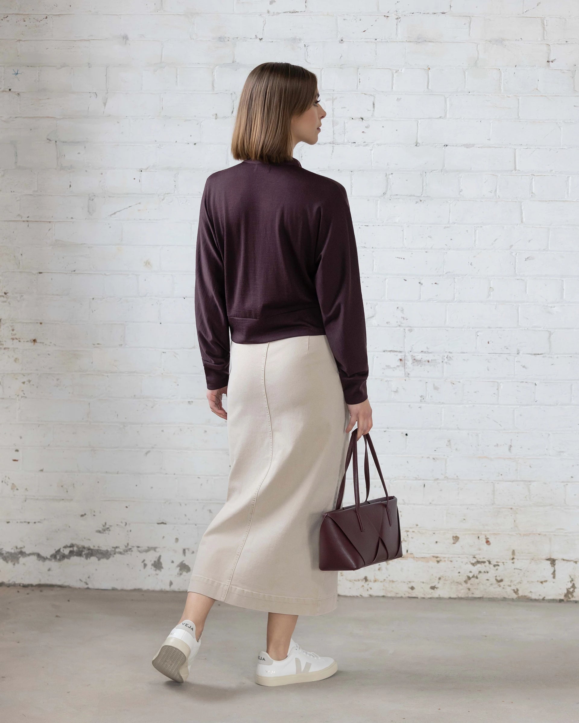 A woman faces away, wearing a long beige skirt, white sneakers, and the Vassalli Nina Merino Cardi in Berry. She holds a maroon handbag and stands against a white brick wall background.