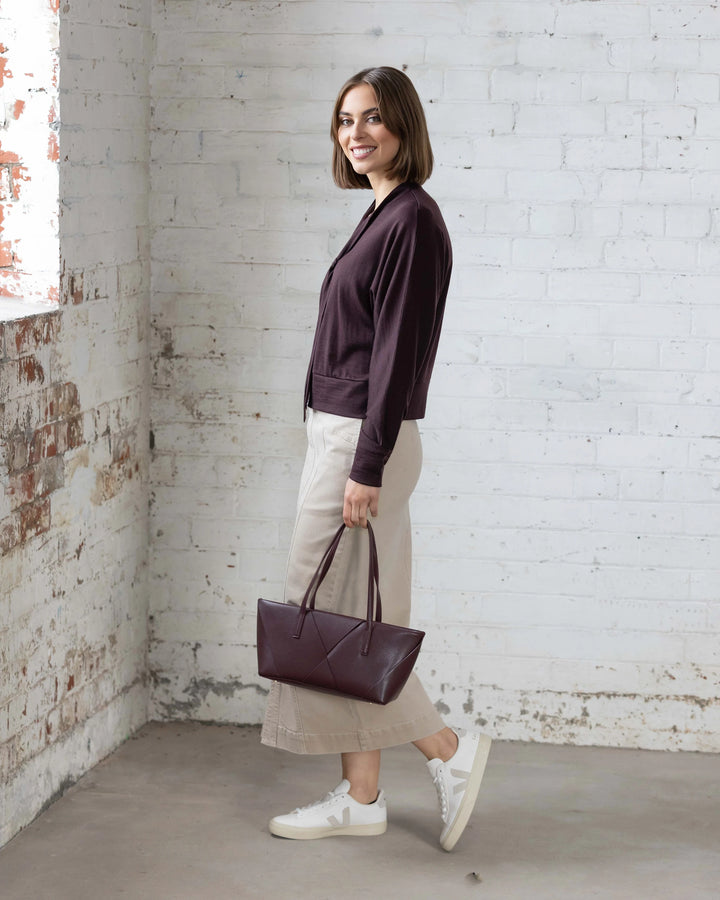 A woman with a brown bob smiles sideways, wearing the Vassalli Nina Merino Cardi in Berry, paired with light beige pants, white sneakers, and holding a matching dark purple handbag against a white brick wall.