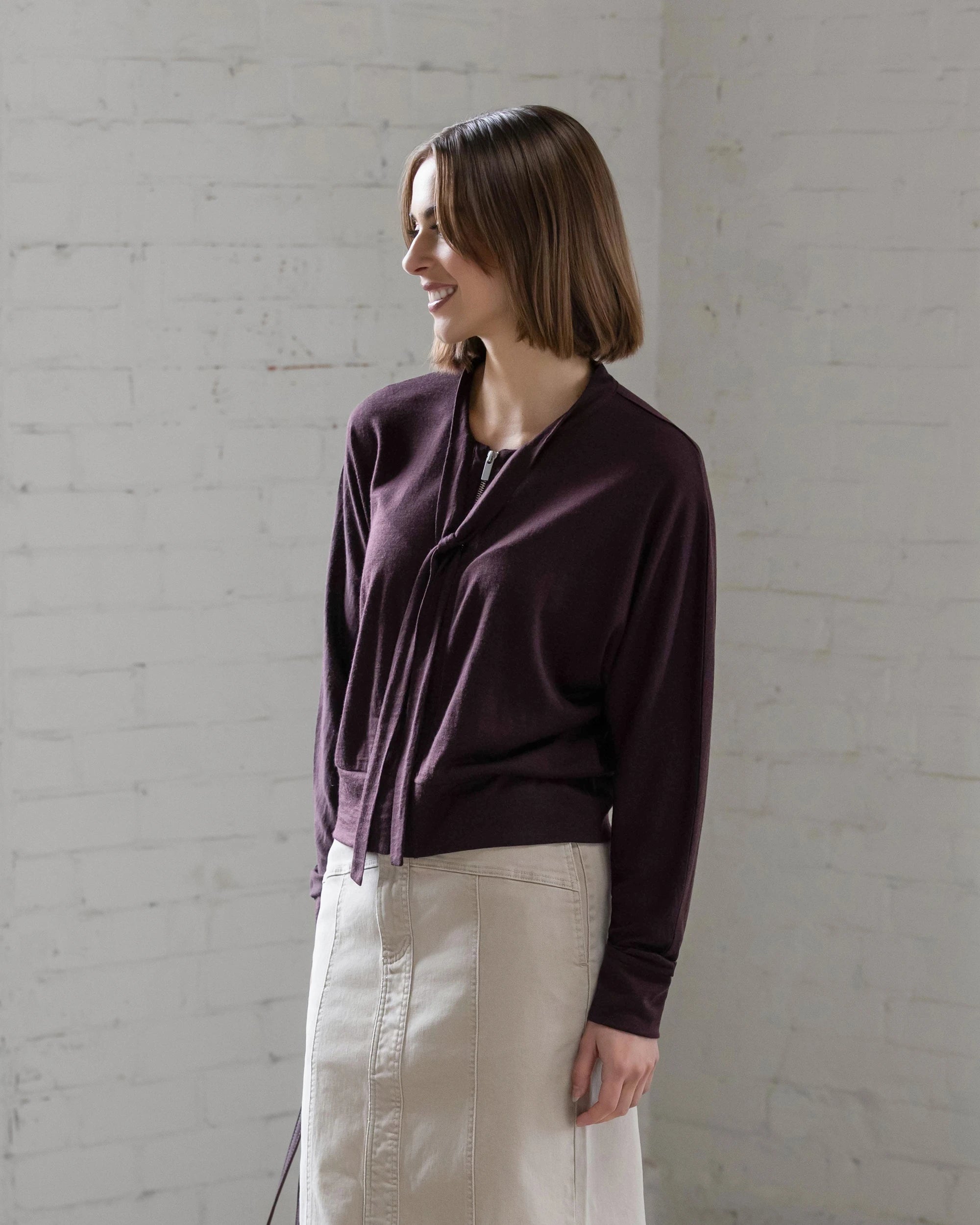 A woman with a straight, shoulder-length bob smiles against a white brick wall, wearing the Vassalli Nina Merino Cardi in Berry paired with a light beige skirt.