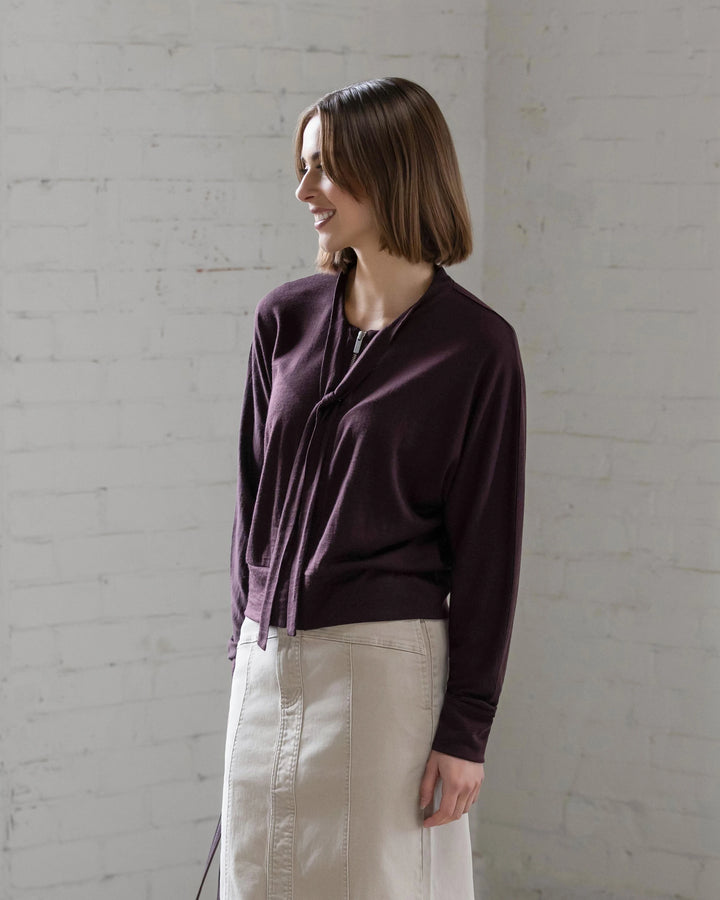 A woman with a straight, shoulder-length bob smiles against a white brick wall, wearing the Vassalli Nina Merino Cardi in Berry paired with a light beige skirt.