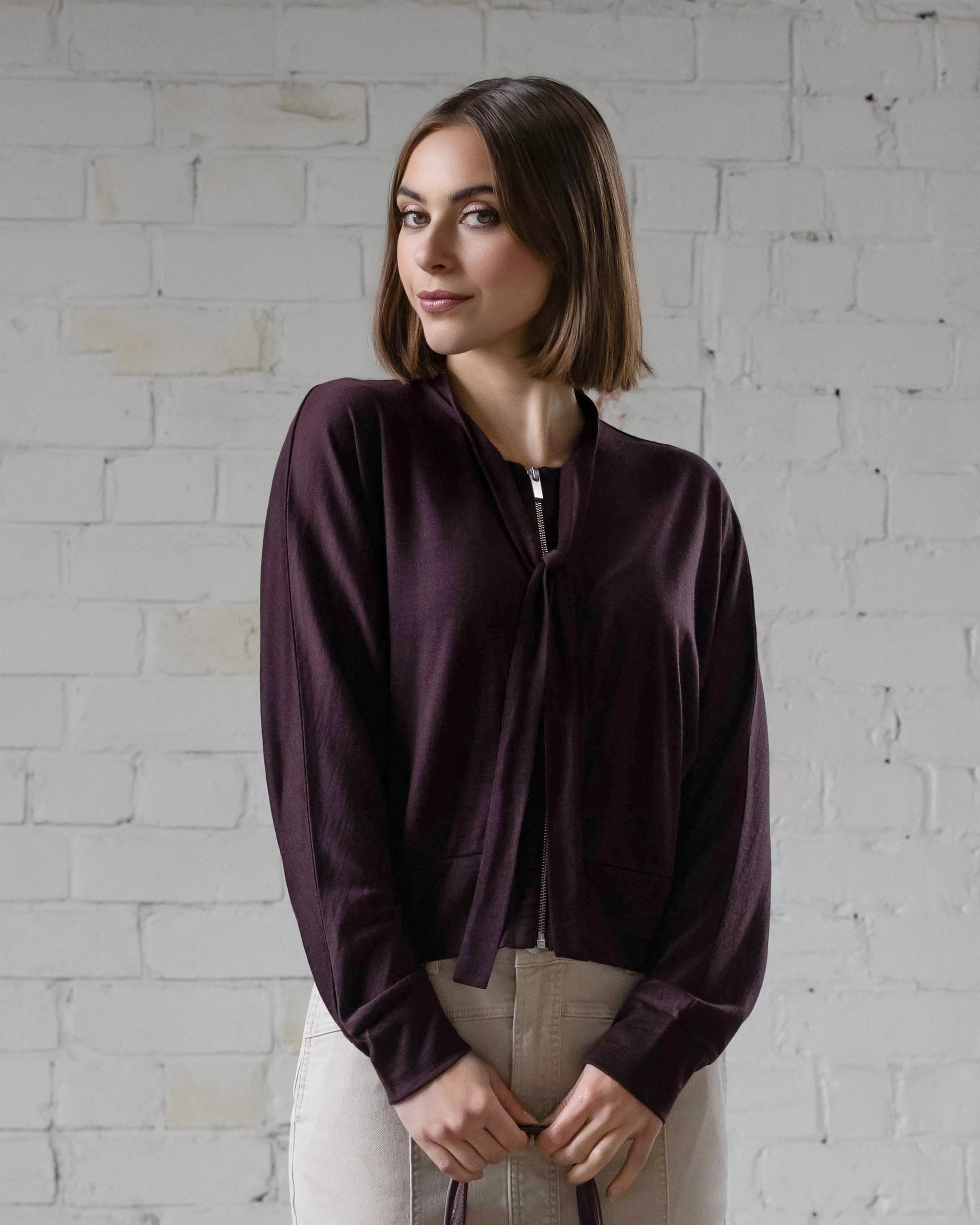A woman with straight brown hair in a chin-length bob wears the Vassalli Nina Merino Cardi in Berry over a dark long-sleeve top and light pants, standing before a white brick wall, smiling subtly and gazing slightly to the side.