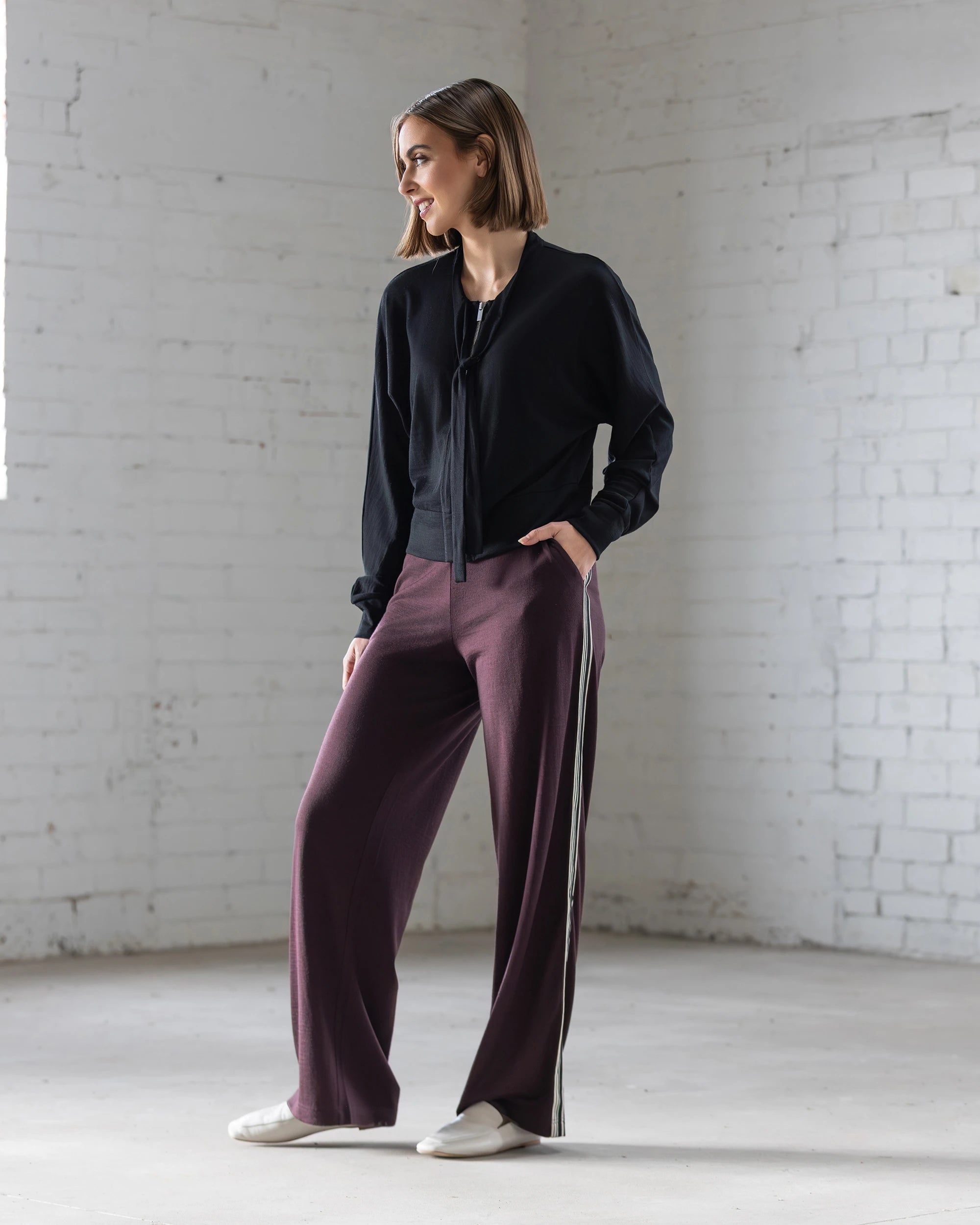 A woman with short hair stands indoors against a white brick wall, wearing the Vassalli Nina Merino Cardi in black with batwing sleeves, wide-legged maroon pants with side stripes, and white shoes. She looks to the side with one hand in her pocket.