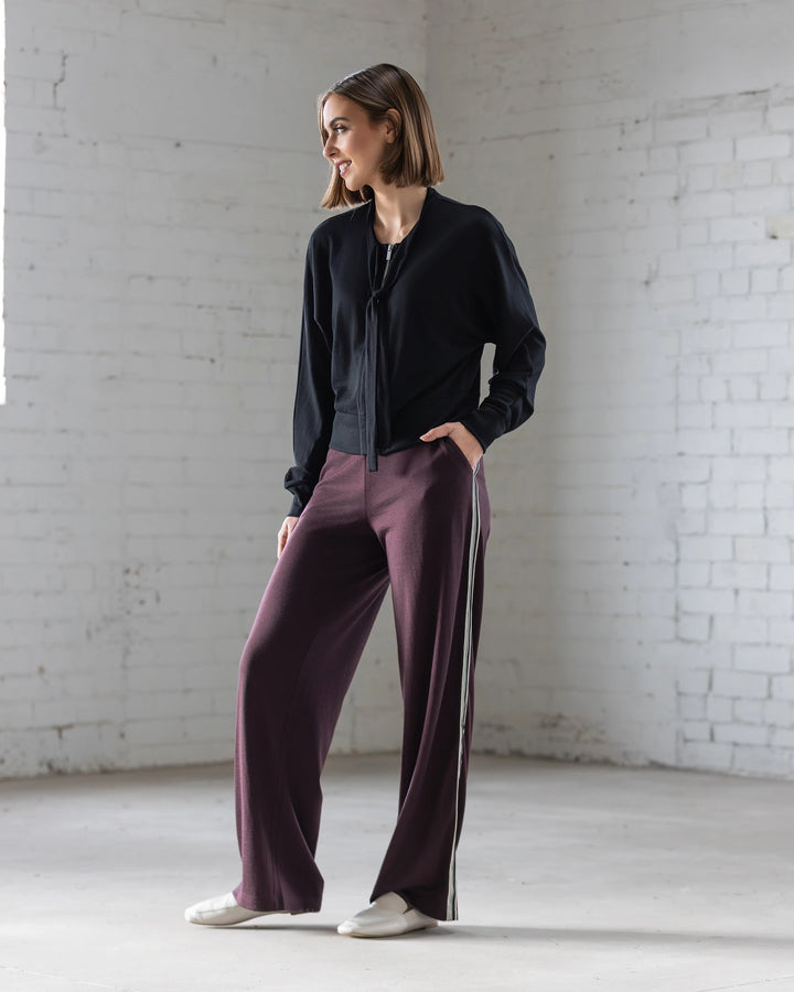 A woman with short hair stands indoors against a white brick wall, wearing the Vassalli Nina Merino Cardi in black with batwing sleeves, wide-legged maroon pants with side stripes, and white shoes. She looks to the side with one hand in her pocket.