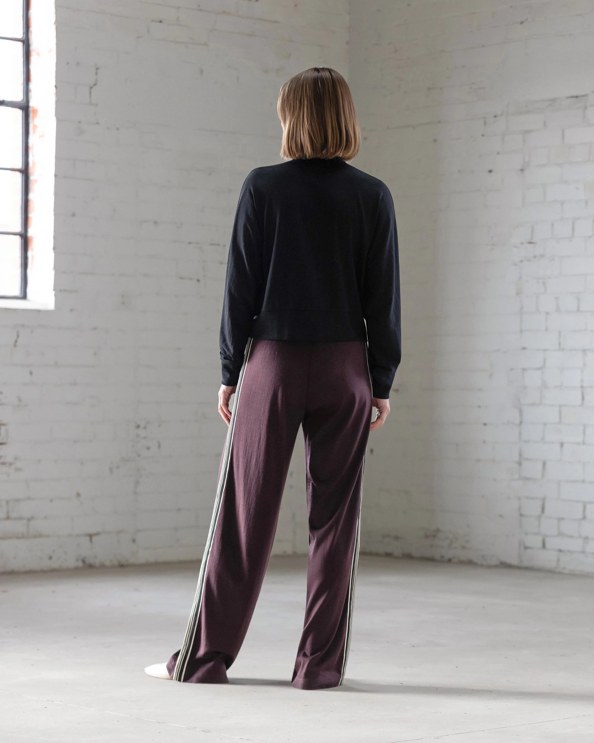 A person with short hair stands indoors facing a white brick wall, wearing the Vassalli Nina Merino Cardi in black and loose maroon pants with side stripes, as light streams in from a window on the left.
