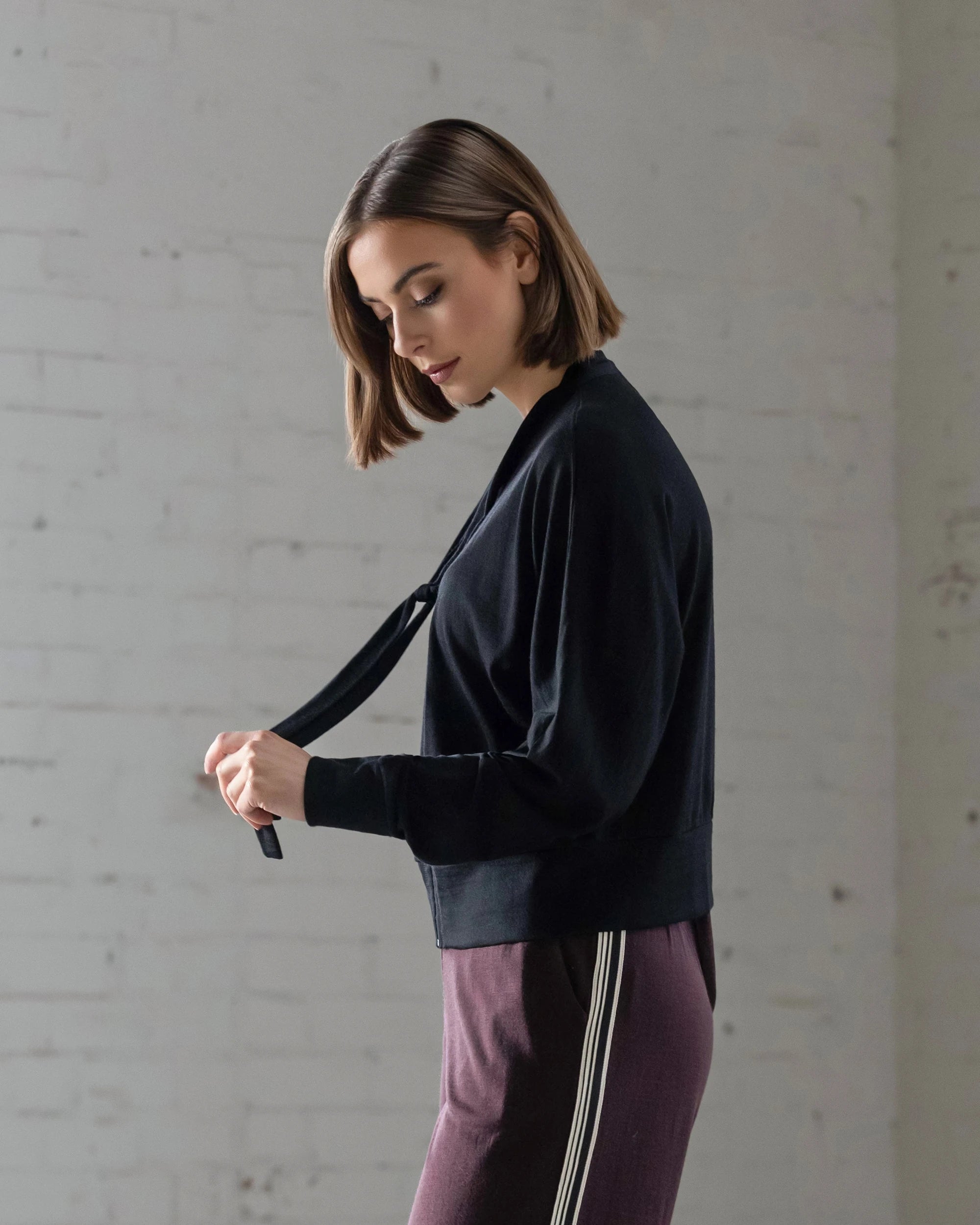 A woman with shoulder-length brown hair stands sideways in front of a white brick wall, wearing the Vassalli Nina Merino Cardi in black and maroon pants with white stripes, looking down while tying her top.