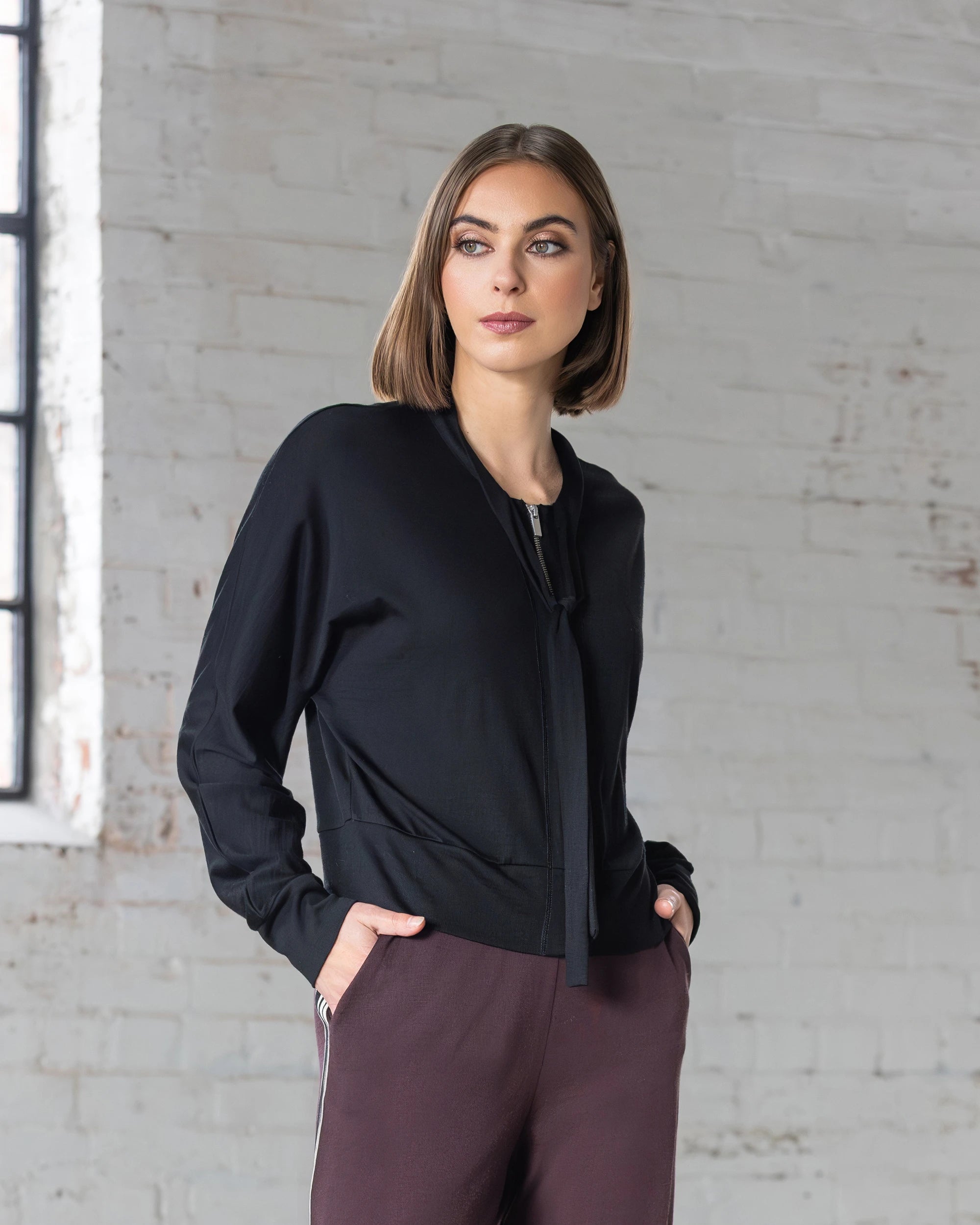 A woman with straight, shoulder-length brown hair wears the Vassalli Nina Merino Cardi in black and maroon pants, standing against a white brick wall with a window on the left. She gazes slightly right with a neutral expression.
