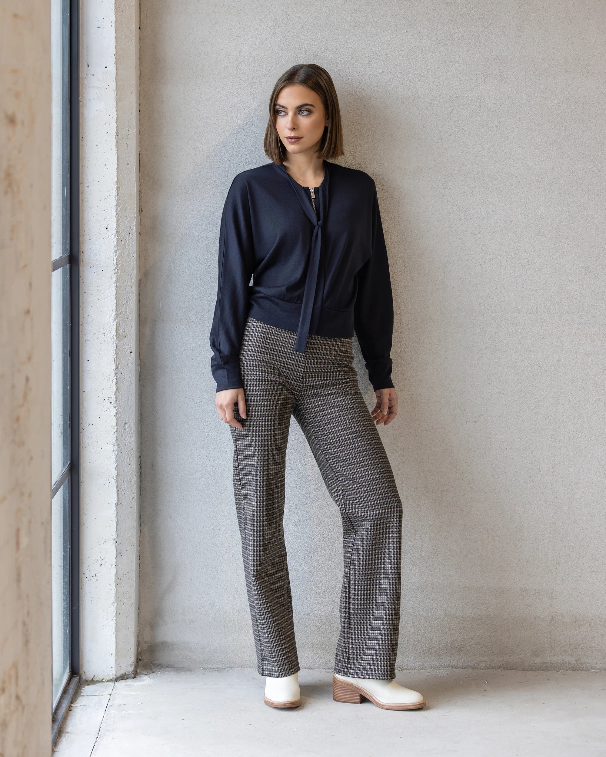 A woman poses indoors by a window against a light gray wall, wearing Vassalli's Nina Merino Cardi in Midnight over a navy tie-neck blouse and plaid high-waisted trousers, finished with cream ankle boots.