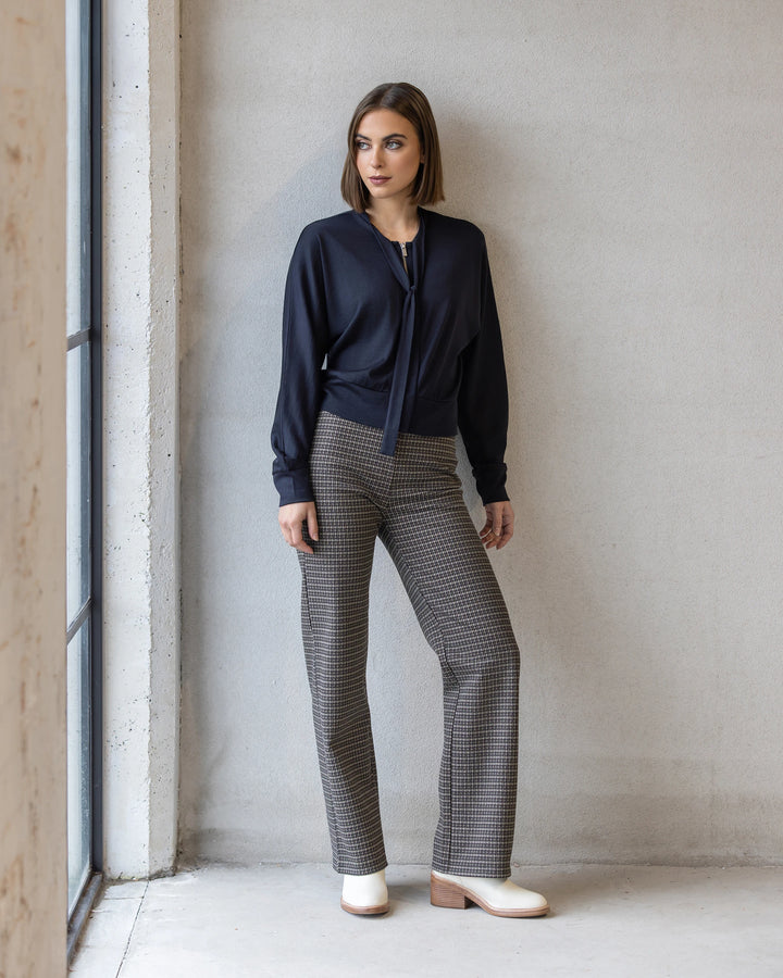 A woman poses indoors by a window against a light gray wall, wearing Vassalli's Nina Merino Cardi in Midnight over a navy tie-neck blouse and plaid high-waisted trousers, finished with cream ankle boots.