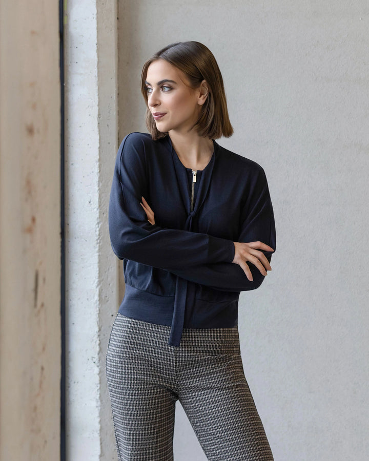 A woman with a bob haircut wears the Vassalli Nina Merino Cardi in Midnight, standing indoors by a concrete wall and window with arms crossed, looking to the side.