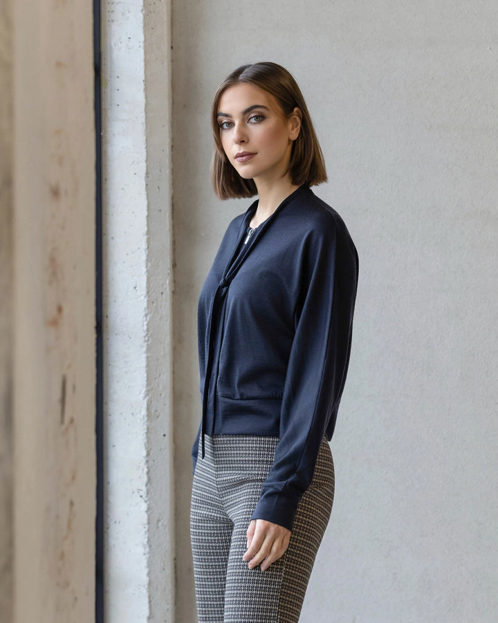 A woman with straight brown hair stands indoors near a white wall, looking at the camera. She is wearing the Vassalli Nina Merino Cardi in Midnight and plaid pants.