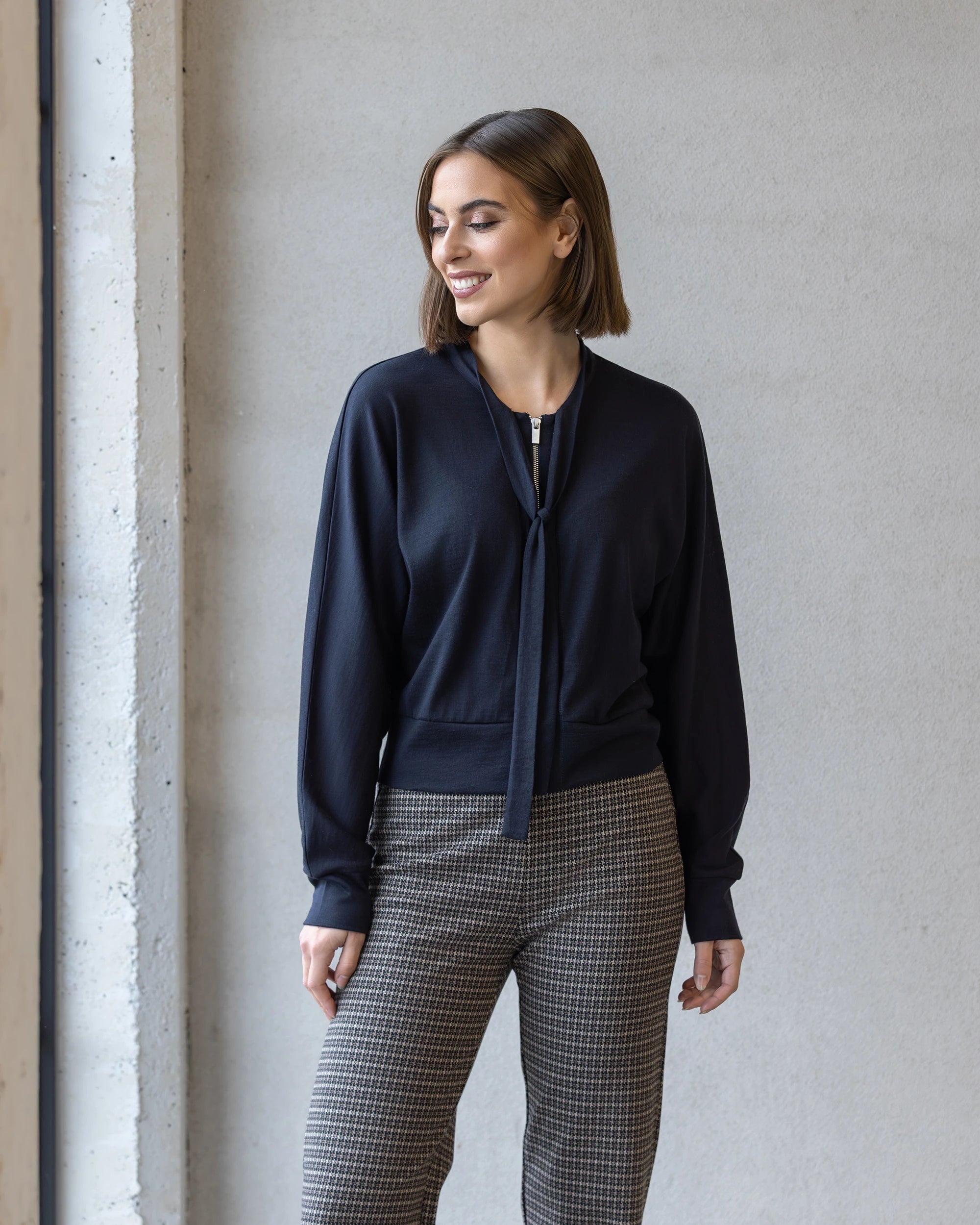 A woman with short brown hair wears the Vassalli Nina Merino Cardi in Midnight, smiling as she stands indoors by a window and looks to the side.