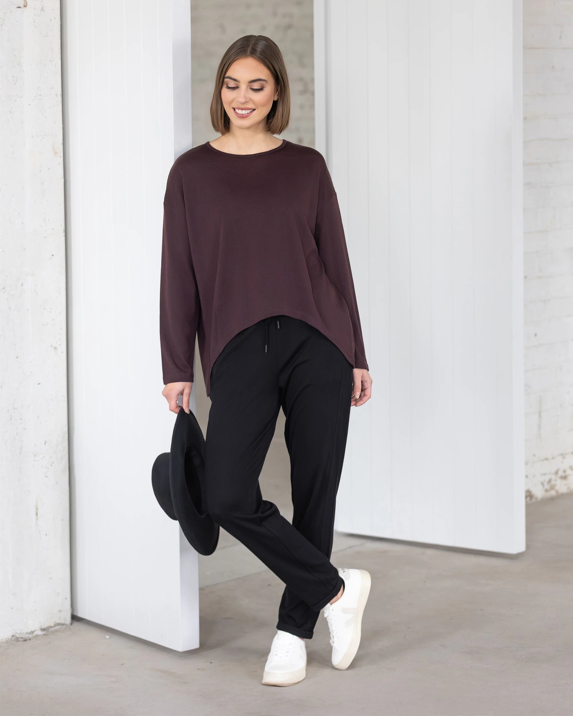 A woman wearing the Vassalli Quinn Merino Top in Berry, paired with black pants and white sneakers, stands indoors near a white wall and door, smiling and holding a black hat by her side.