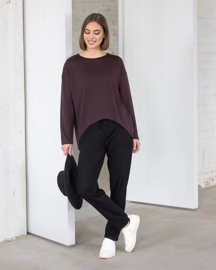 A woman wearing the Vassalli Quinn Merino Top in Berry, paired with black pants and white sneakers, stands indoors near a white wall and door, smiling and holding a black hat by her side.