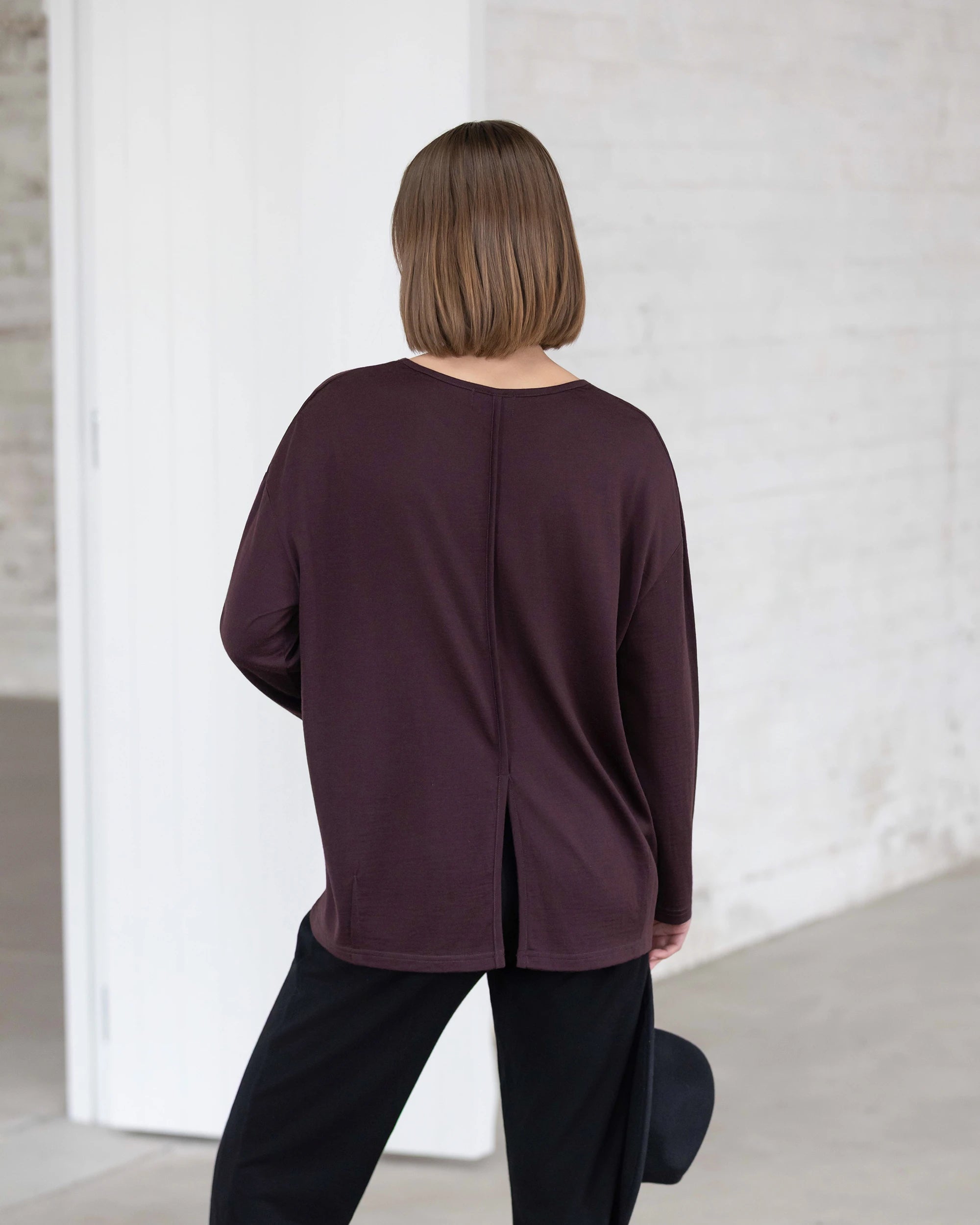 A person with straight, shoulder-length brown hair stands indoors, facing away from the camera, wearing the Vassalli Quinn Merino Top in berry and black pants, holding a hat in one hand.