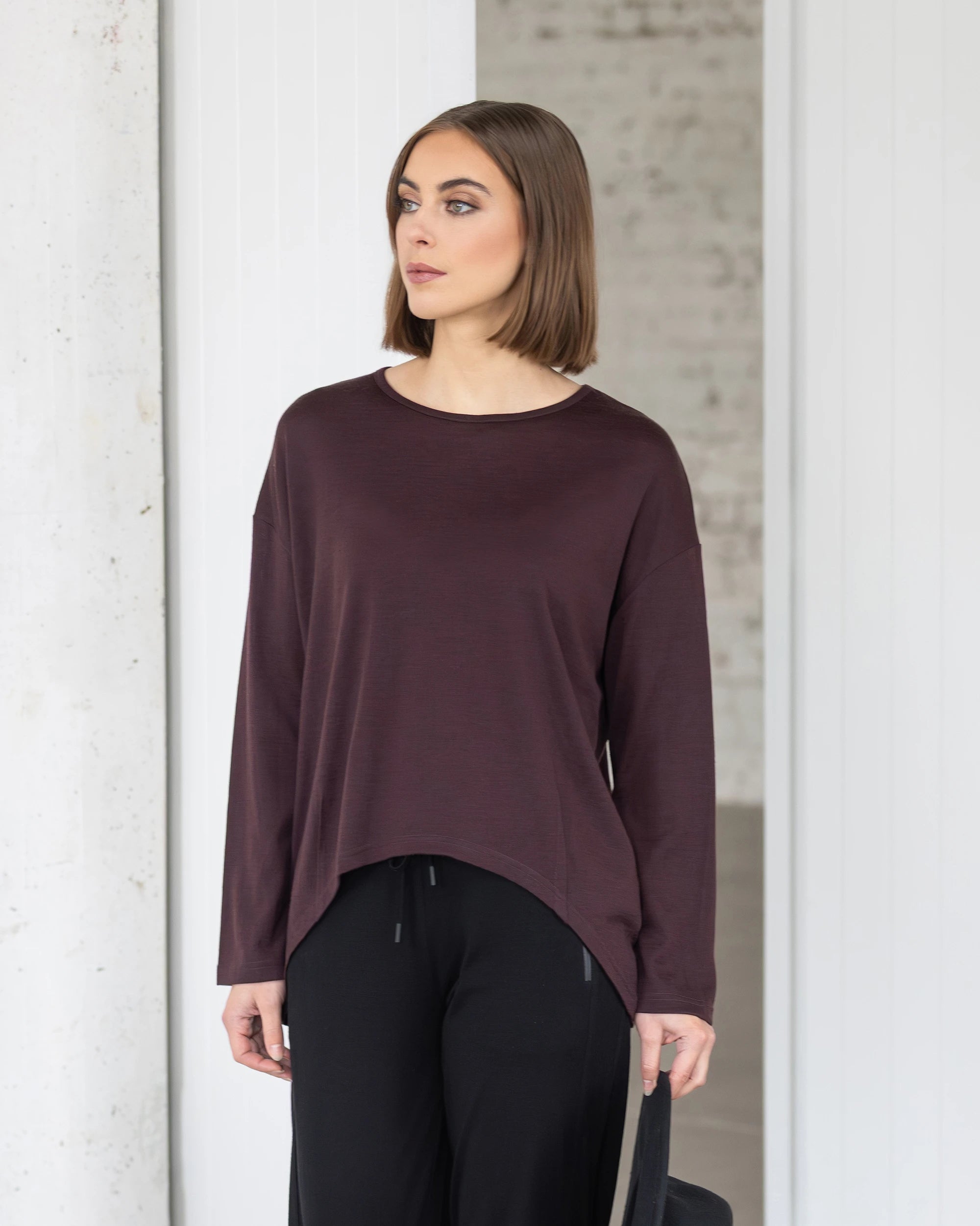 A woman with a bob haircut wears the Vassalli Quinn Merino Top in berry and black pants, standing indoors against a white and light gray background, looking to her left.