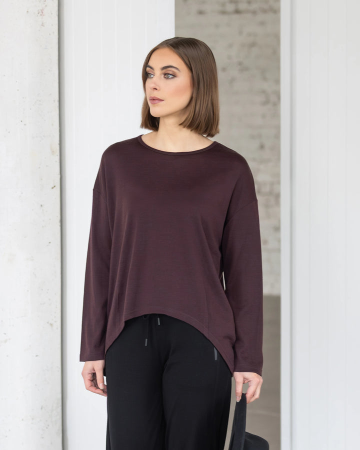 A woman with a bob haircut wears the Vassalli Quinn Merino Top in berry and black pants, standing indoors against a white and light gray background, looking to her left.