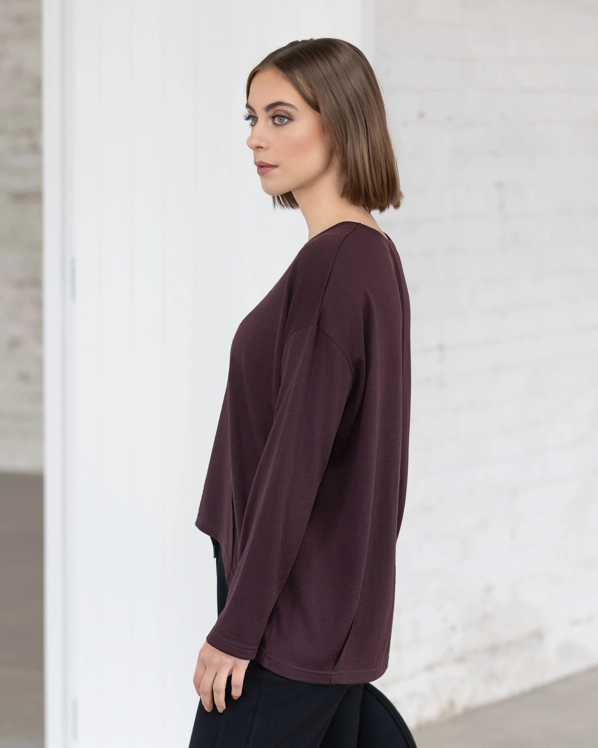 A woman with straight, shoulder-length brown hair stands indoors in profile, wearing the Vassalli Quinn Merino Top in Berry and black pants. The minimal setting features white brick walls and a doorway.